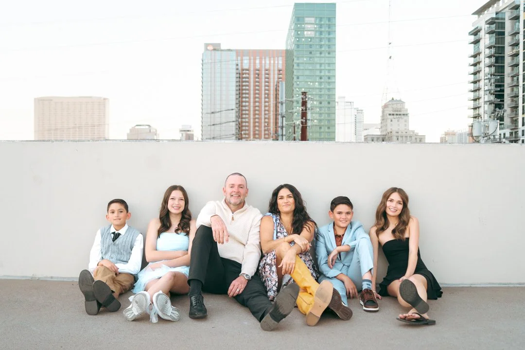 A family of six sitting on the ground in front of a cityscape with tall buildings, smiling at the camera.