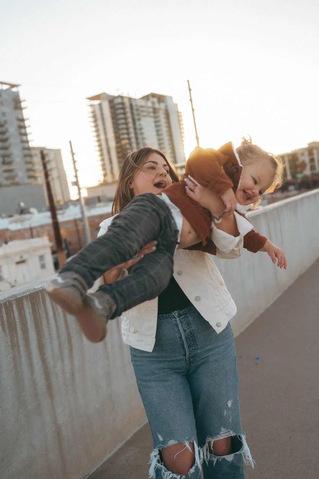 A woman holding a young girl in the air on a city sidewalk during sunset.