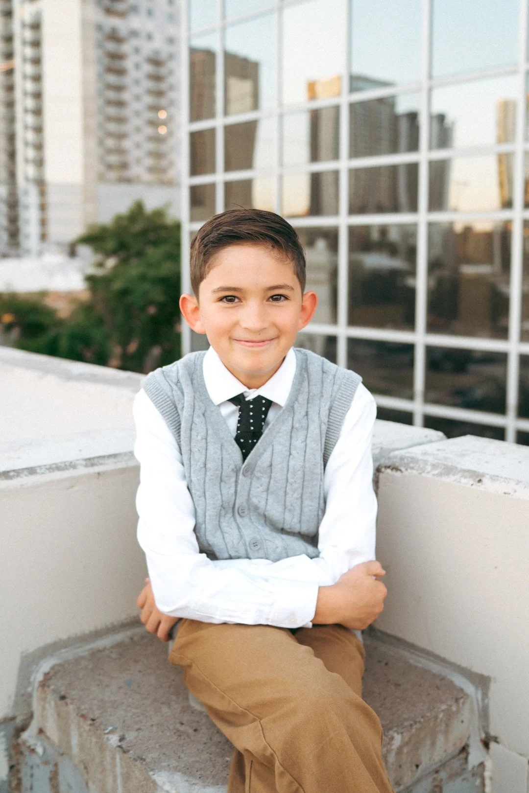 A young boy sitting on a concrete ledge outdoors, smiling at the camera with city buildings and a glass windowed building in the background.