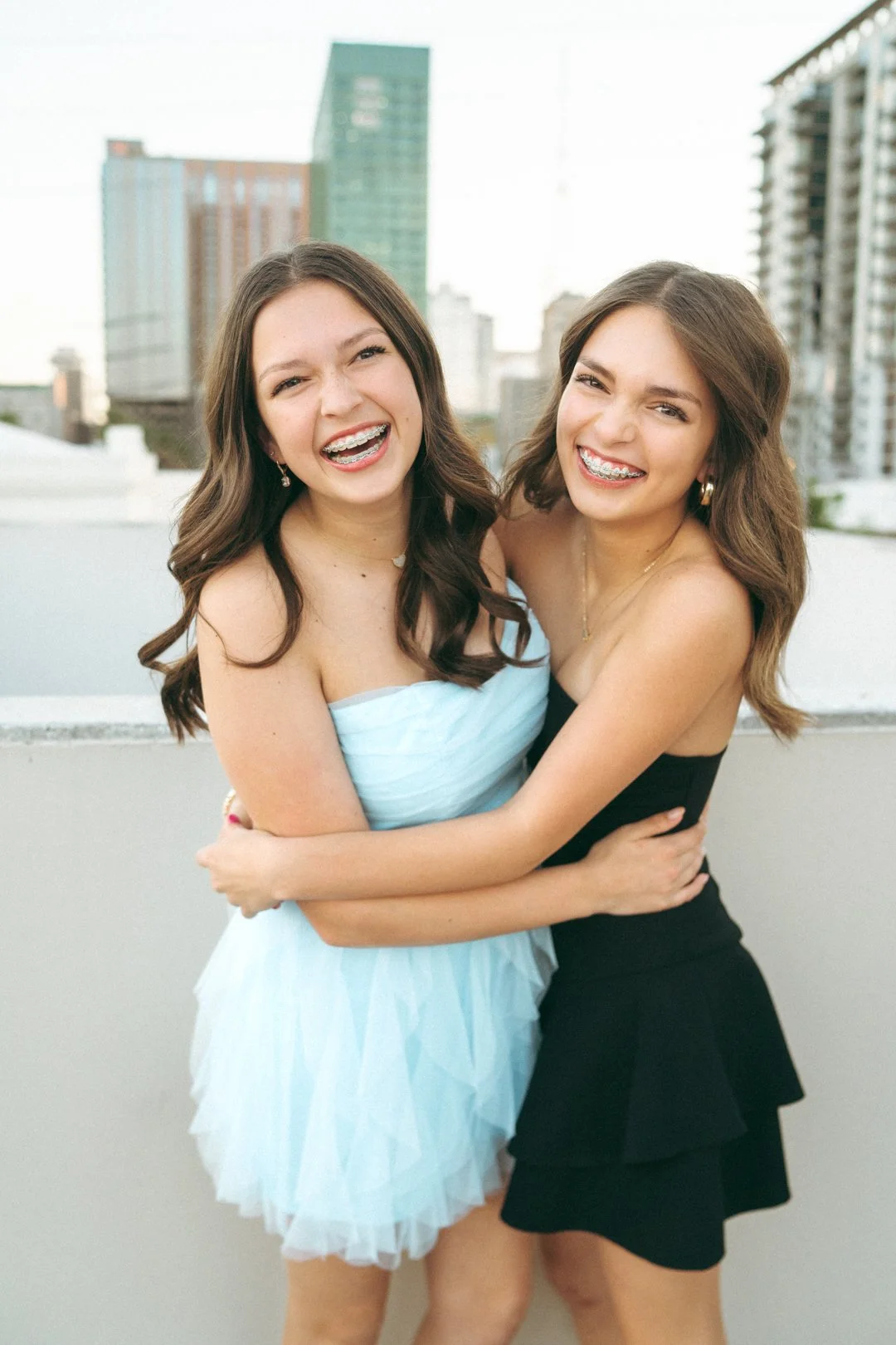 Two young women smiling and hugging each other on a rooftop with city buildings in the background.