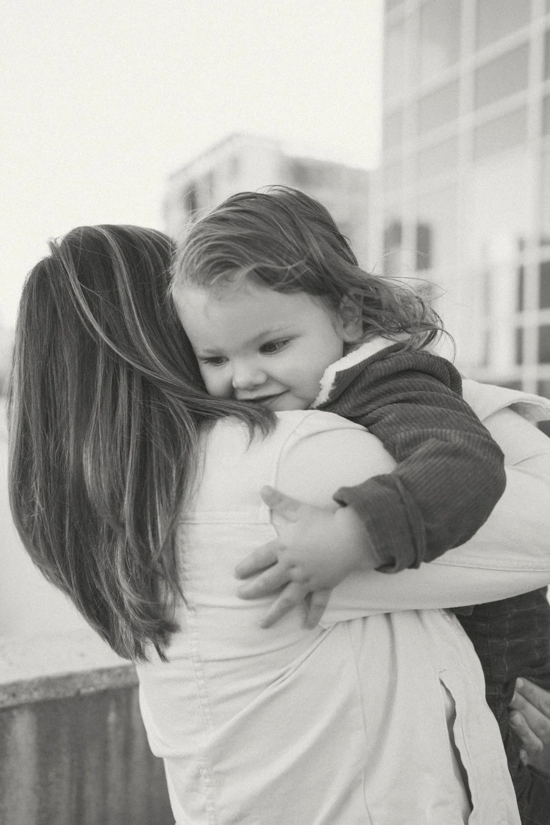 A young child with wet hair hugging a woman with long hair in an outdoor setting, with buildings in the background.
