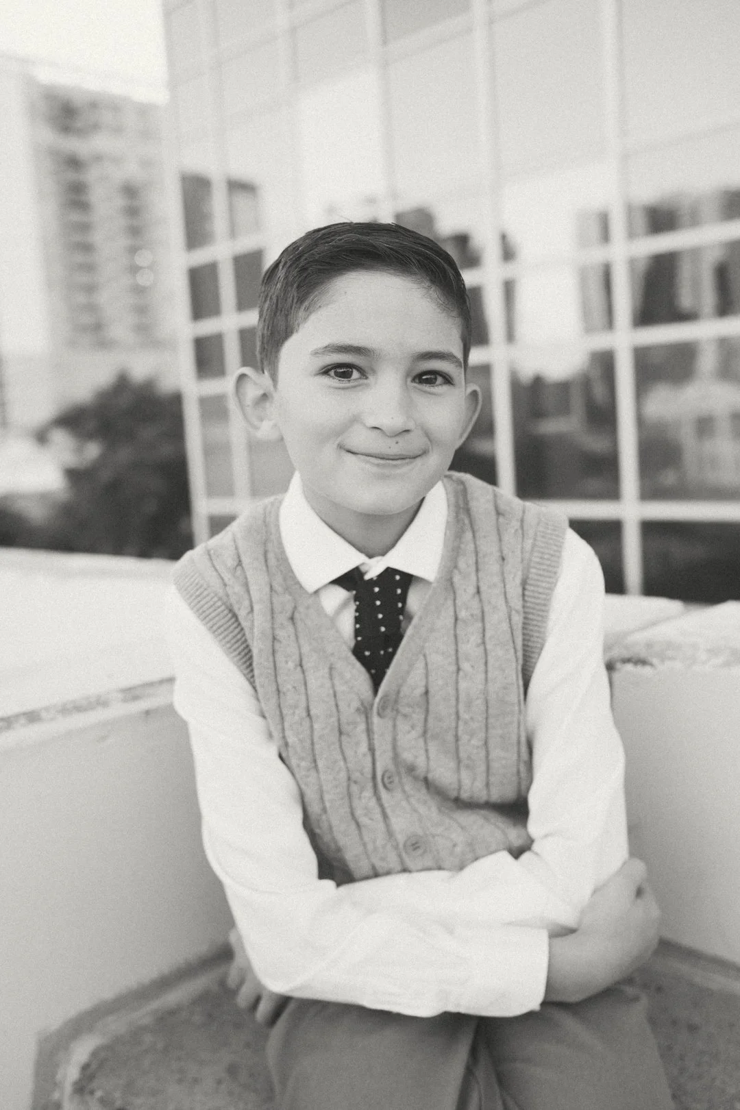 A young boy with short dark hair, wearing a white shirt, polka dot tie, and a knit vest, sitting with arms crossed and smiling at the camera.