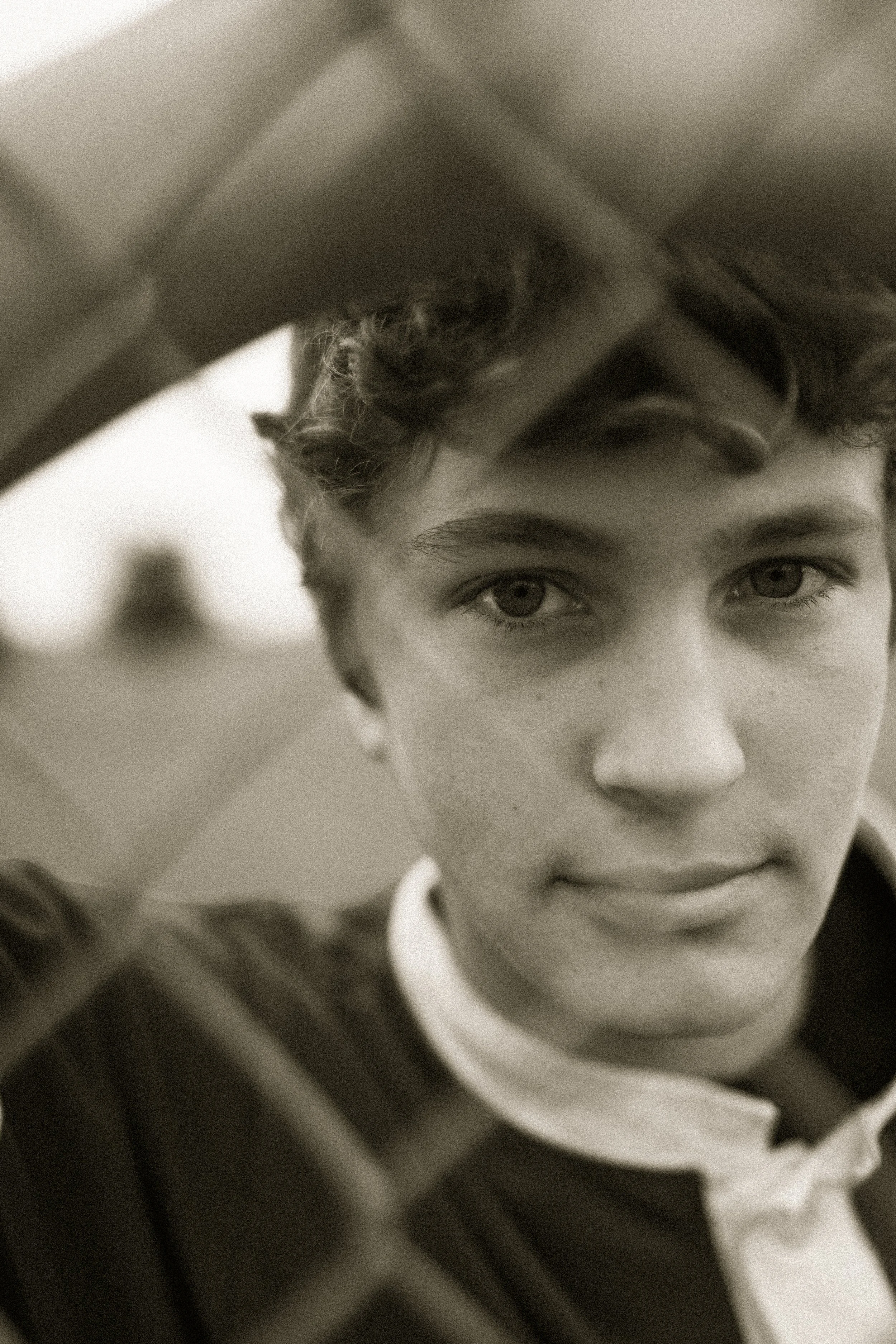 Close-up black-and-white photo of a young man with wavy hair and piercing eyes, partially obscured by window blinds.