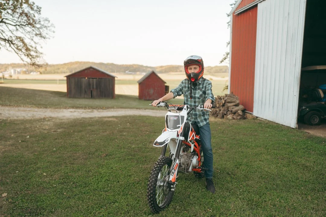 A boy wearing a helmet and plaid shirt sitting on a dirt bike in a rural farm setting with red barns and open fields in the background.