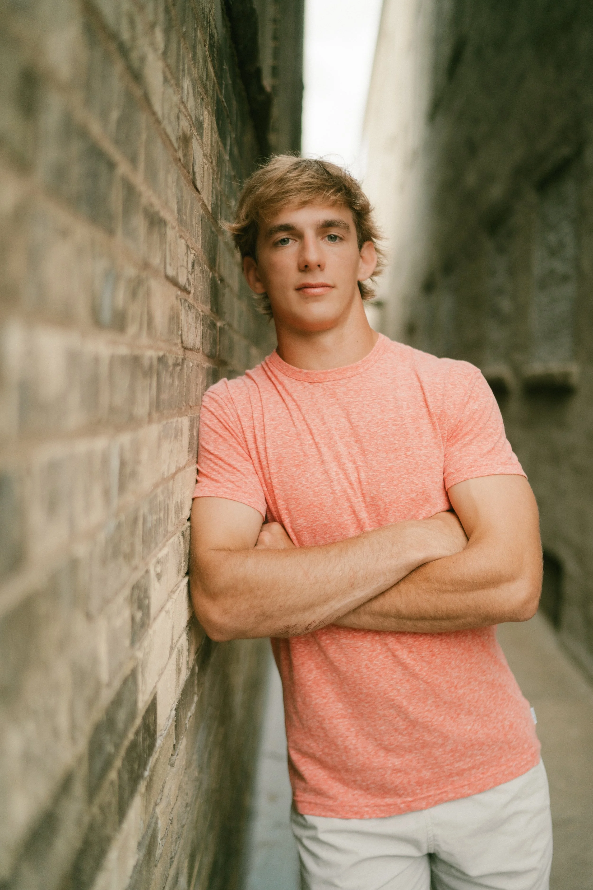 A young man with light brown hair wearing a coral pink t-shirt and cream shorts, crossing his arms, standing in an alleyway with brick and concrete walls.