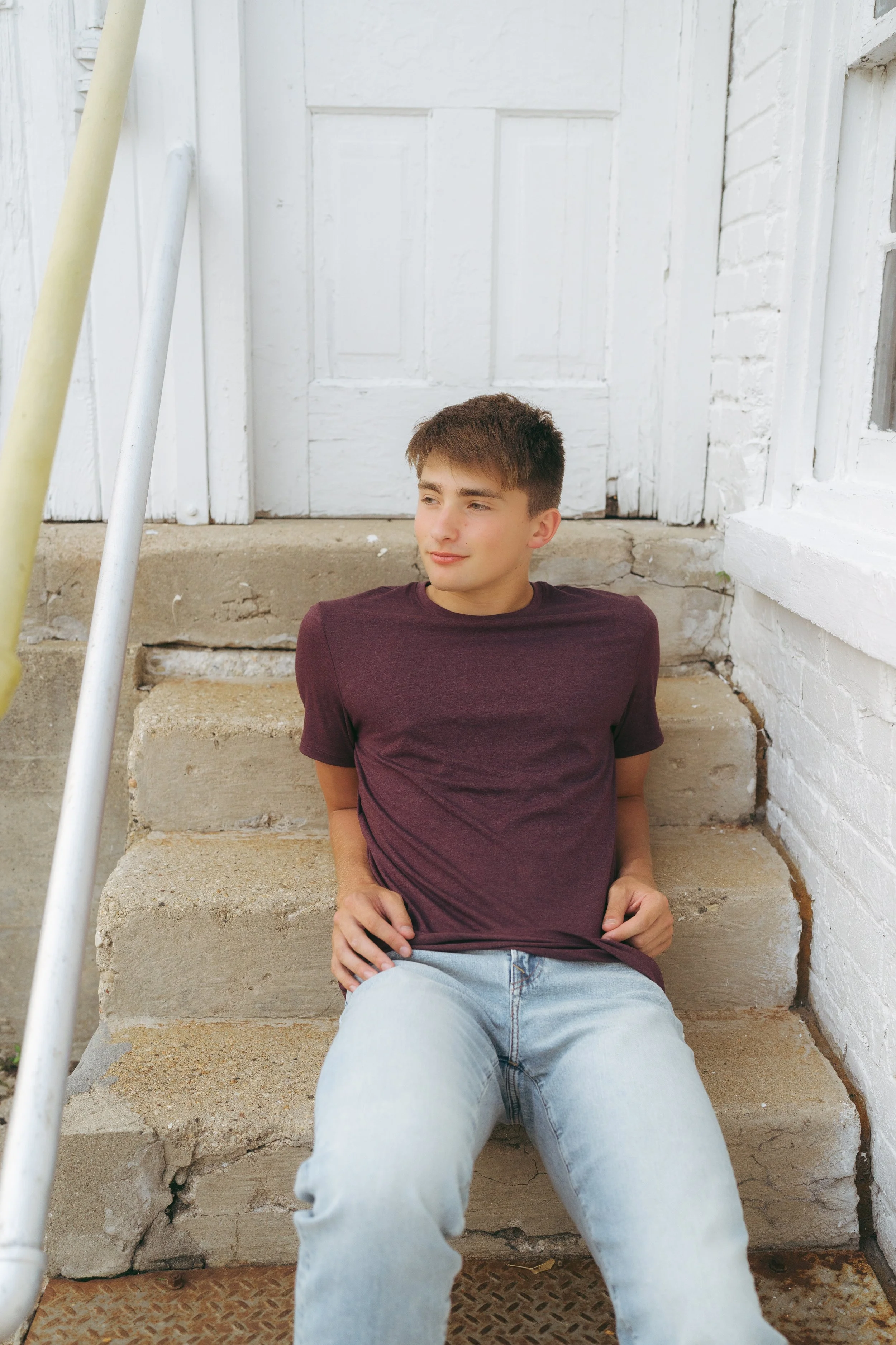 Young man with short brown hair sitting on outdoor concrete steps, wearing a purple t-shirt and light blue jeans, looking to the side against a white brick wall and white door.