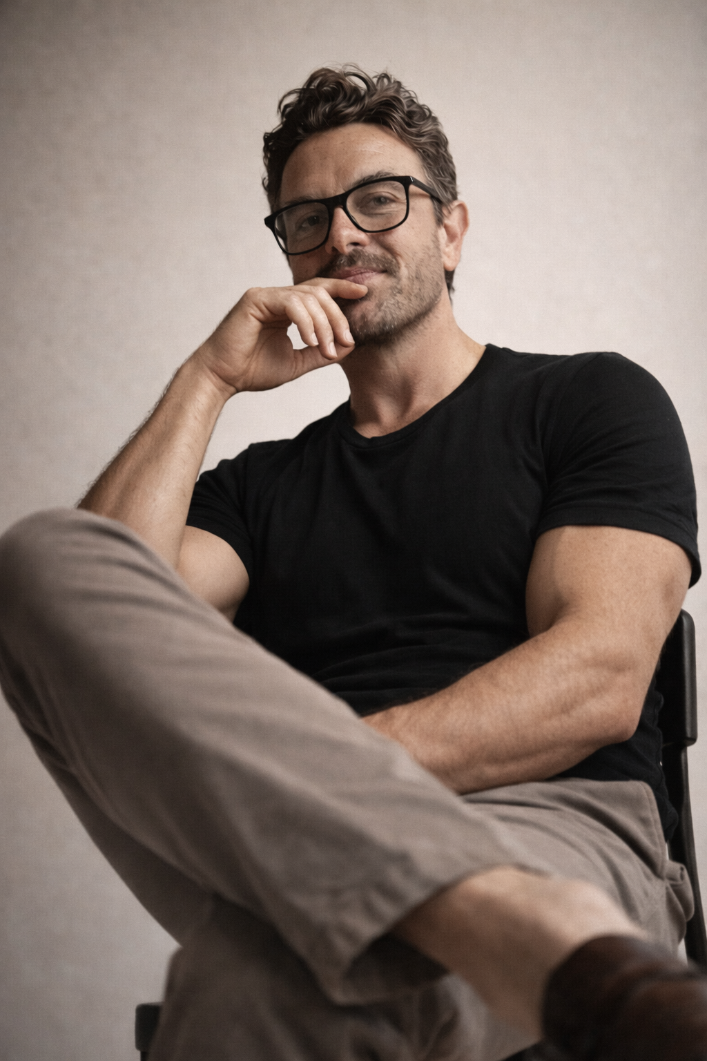 A man with curly brown hair, wearing glasses and a black t-shirt, sits with his right hand near his face, smiling slightly, against a plain wall background.
