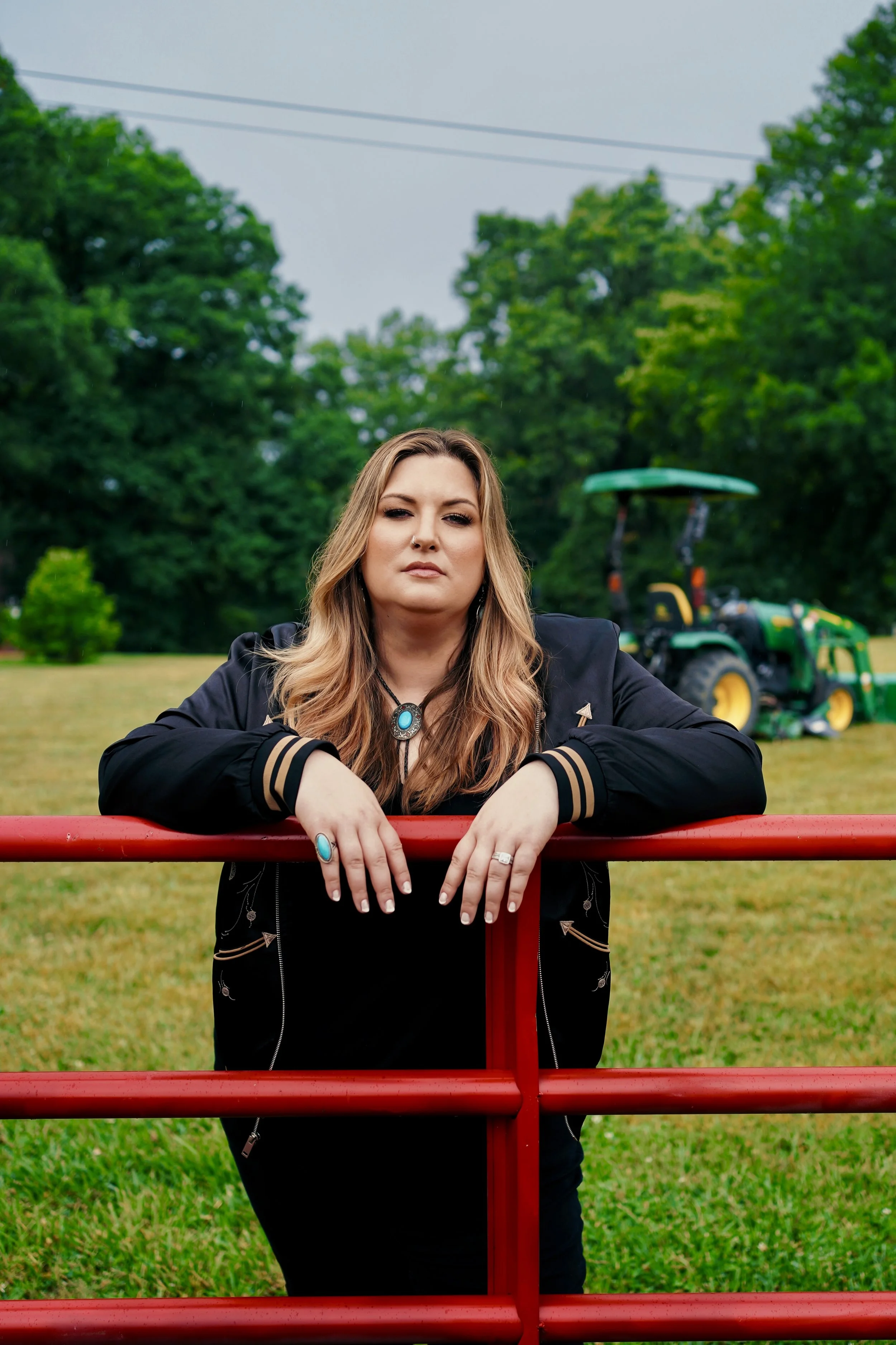 Woman with long blonde hair standing outdoors, leaning on a red fence, dressed in a black jacket with gold accents, wearing jewelry including rings, a necklace with a blue pendant, and earrings, with a green tractor and trees in the background.
