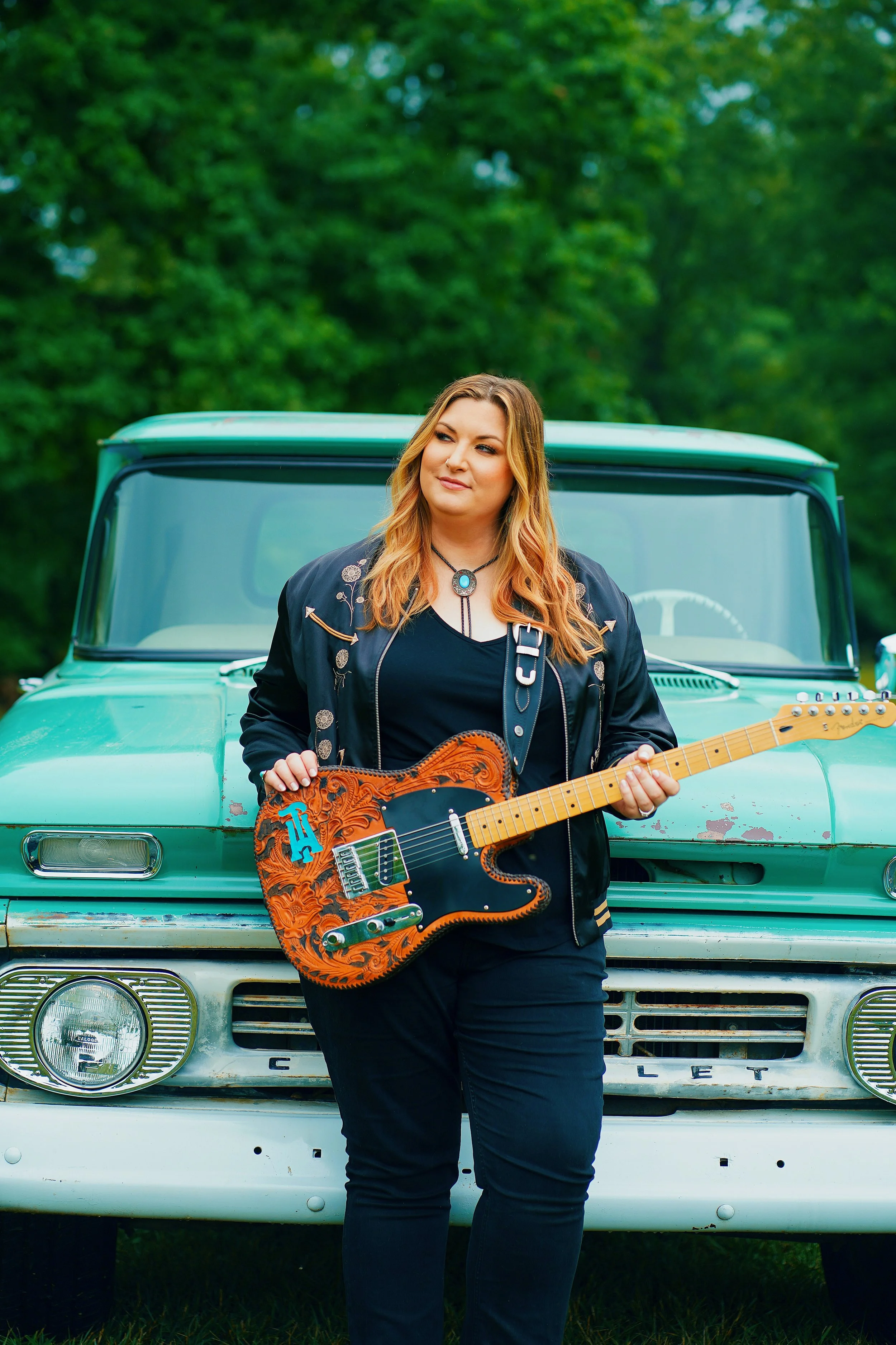 Woman with long red hair wearing a black jacket and necklace, holding an electric guitar, standing in front of a vintage green pickup truck with a forest background.