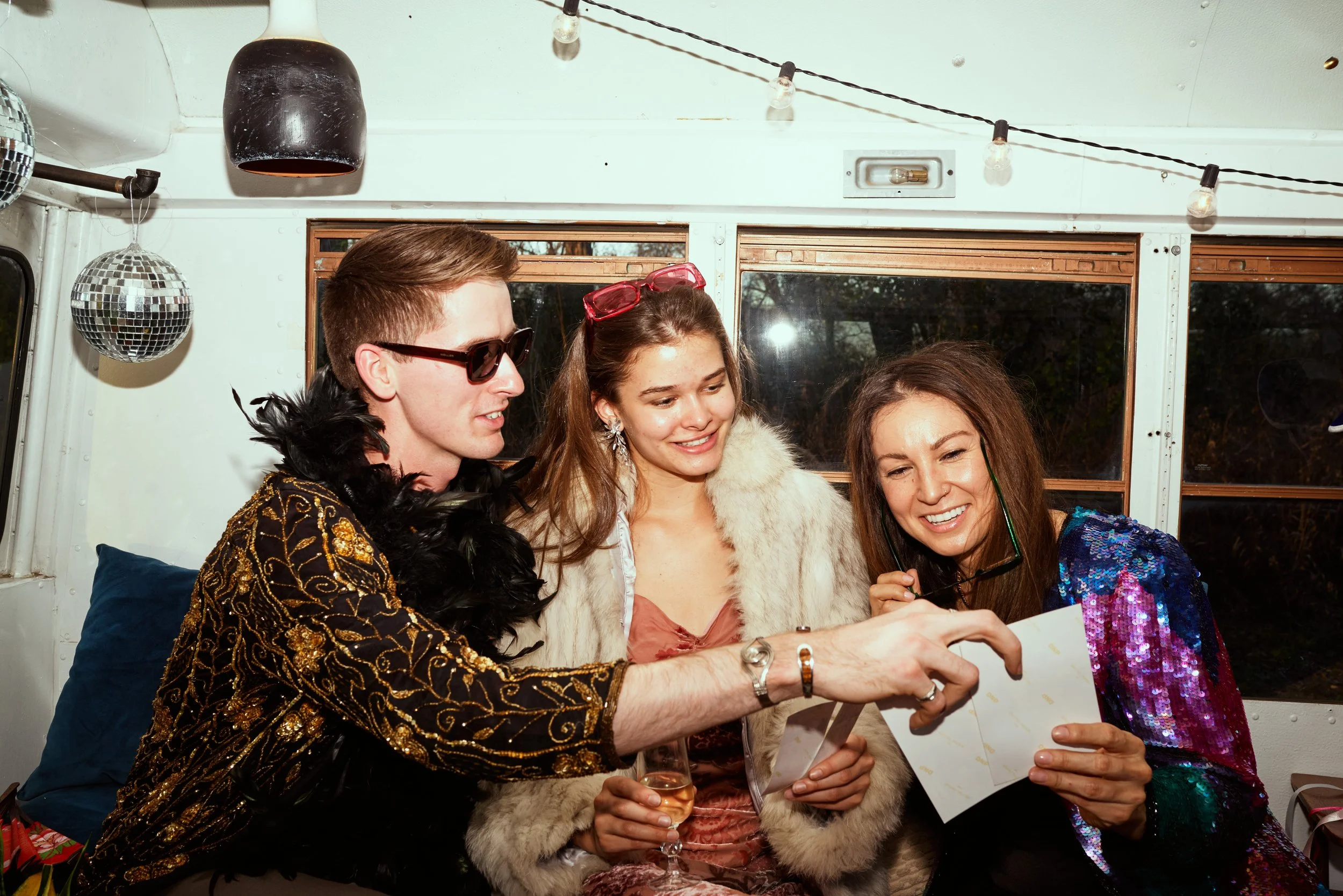 Three people inside a bus wearing party outfits, holding a photo, with disco balls and string lights hanging above.