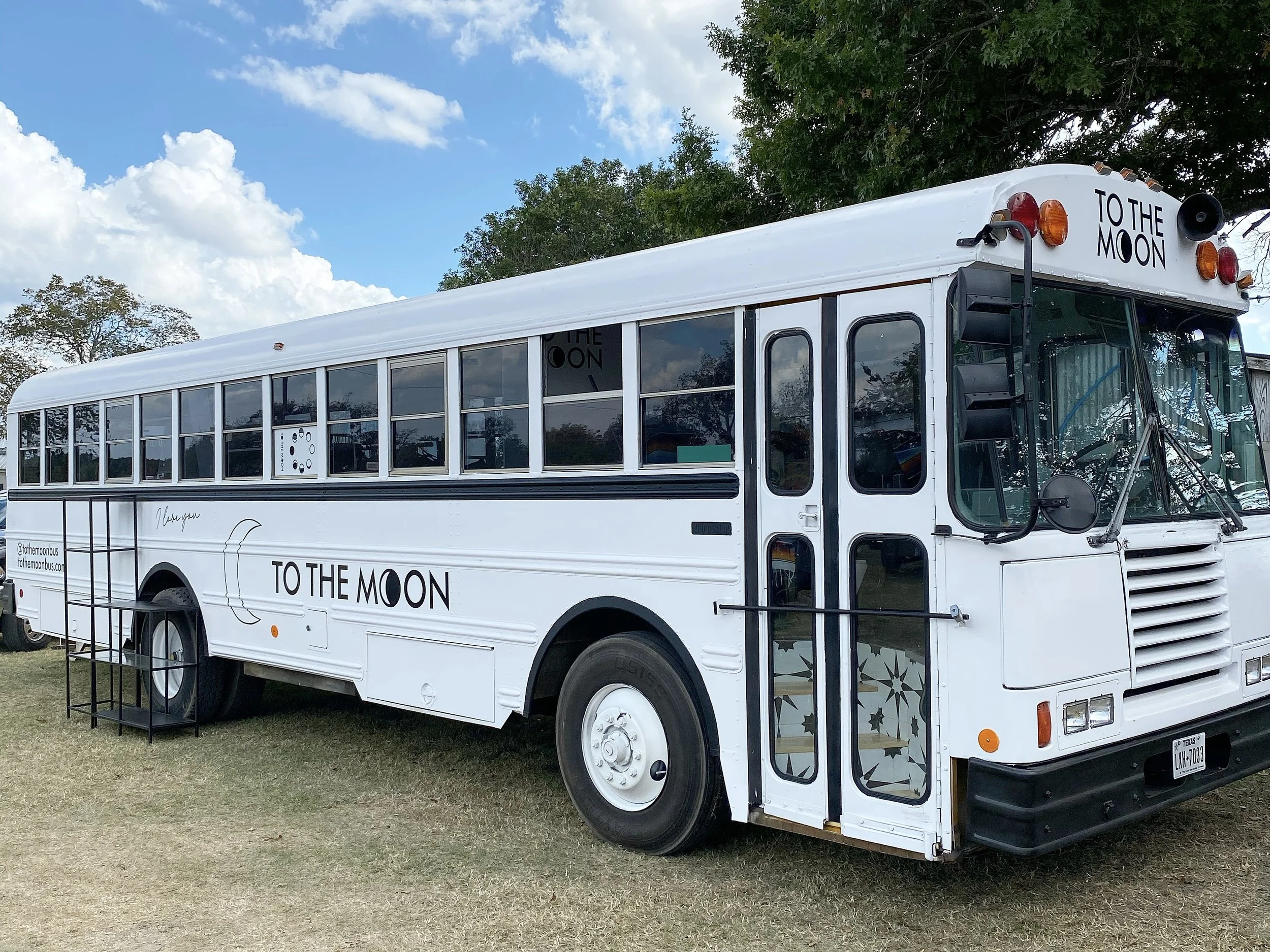 White bus labeled 'To The Moon' parked on grass with steps leading to the entrance, trees and sky in the background.