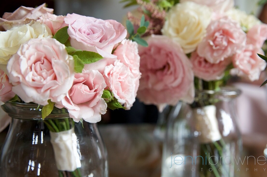 Bridal bouquets containing light pink and cream roses sitting in glass jars. 