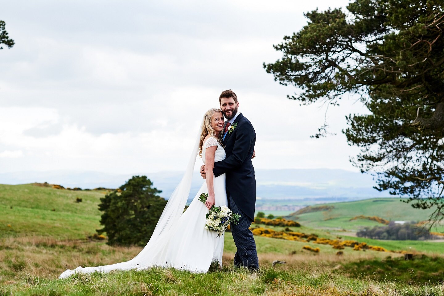 Bride and Groom standing in Scottish hills, bride holding wedding flowers  by her side