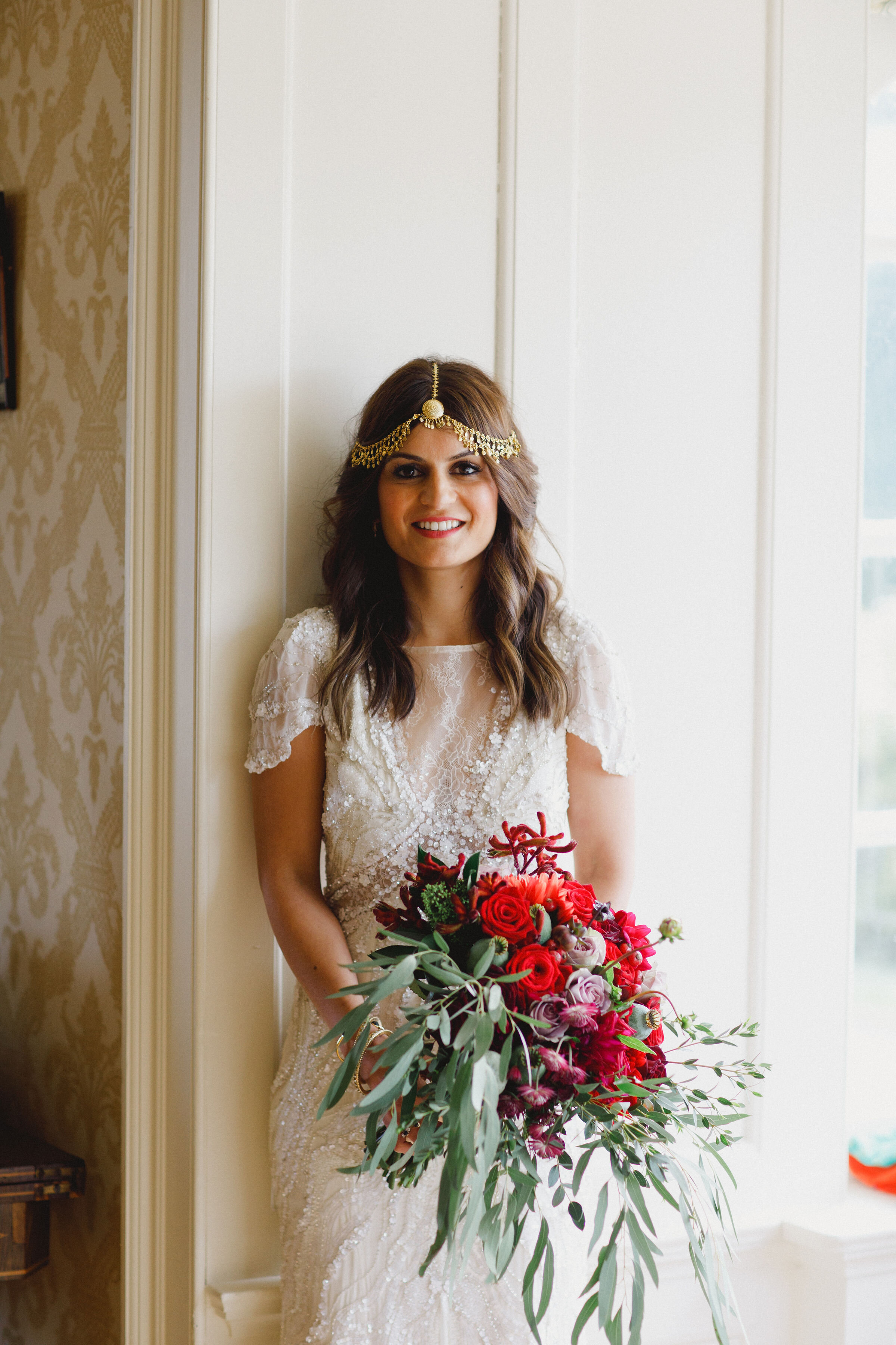 Brides wearing a beautiful beaded gown and holding red and green hand-tied bridal bouquet with lots of foliage.