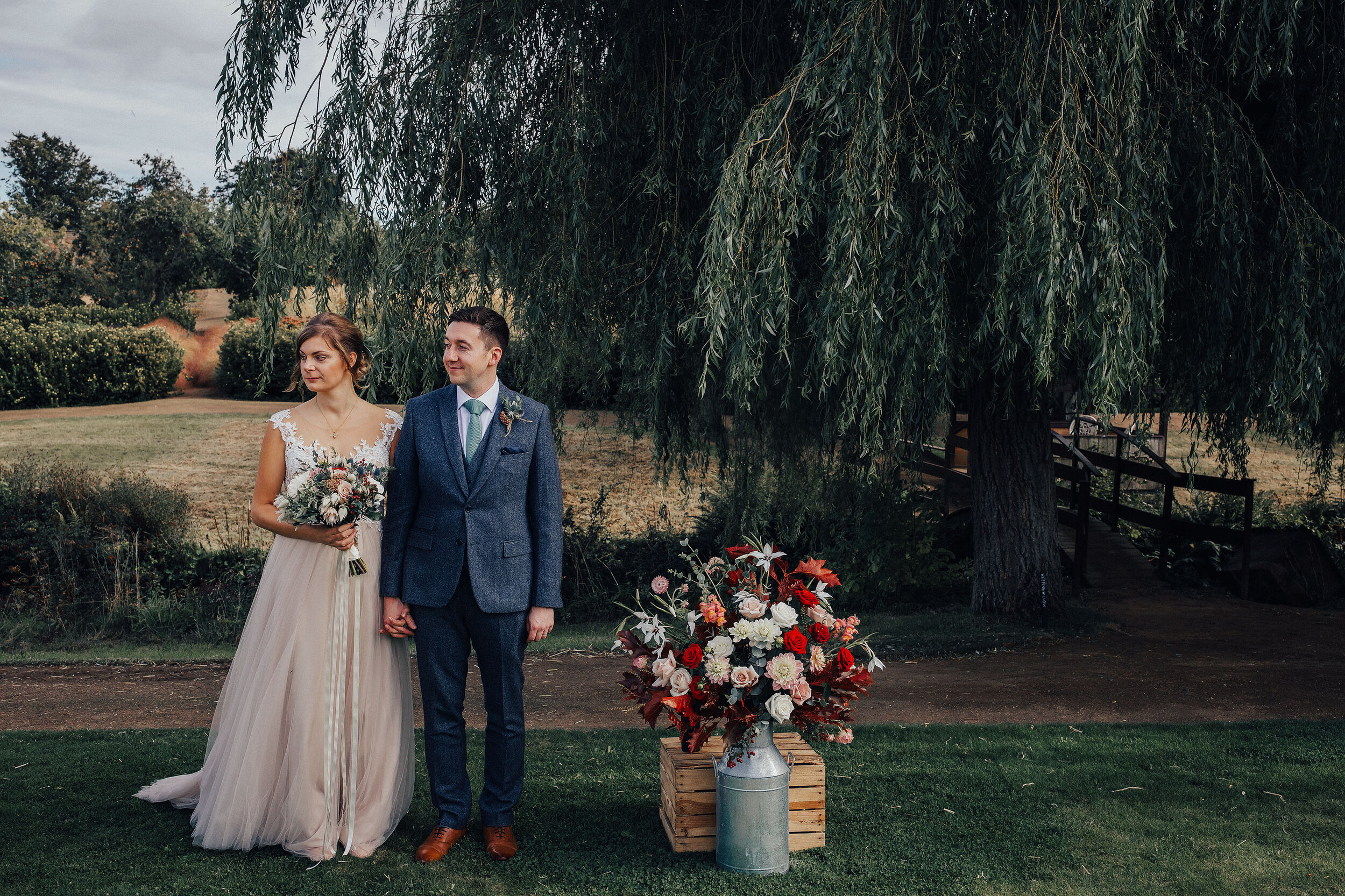 Bride and Groom standing at wedding ceremony with floral arrangement displayed in milk churn 