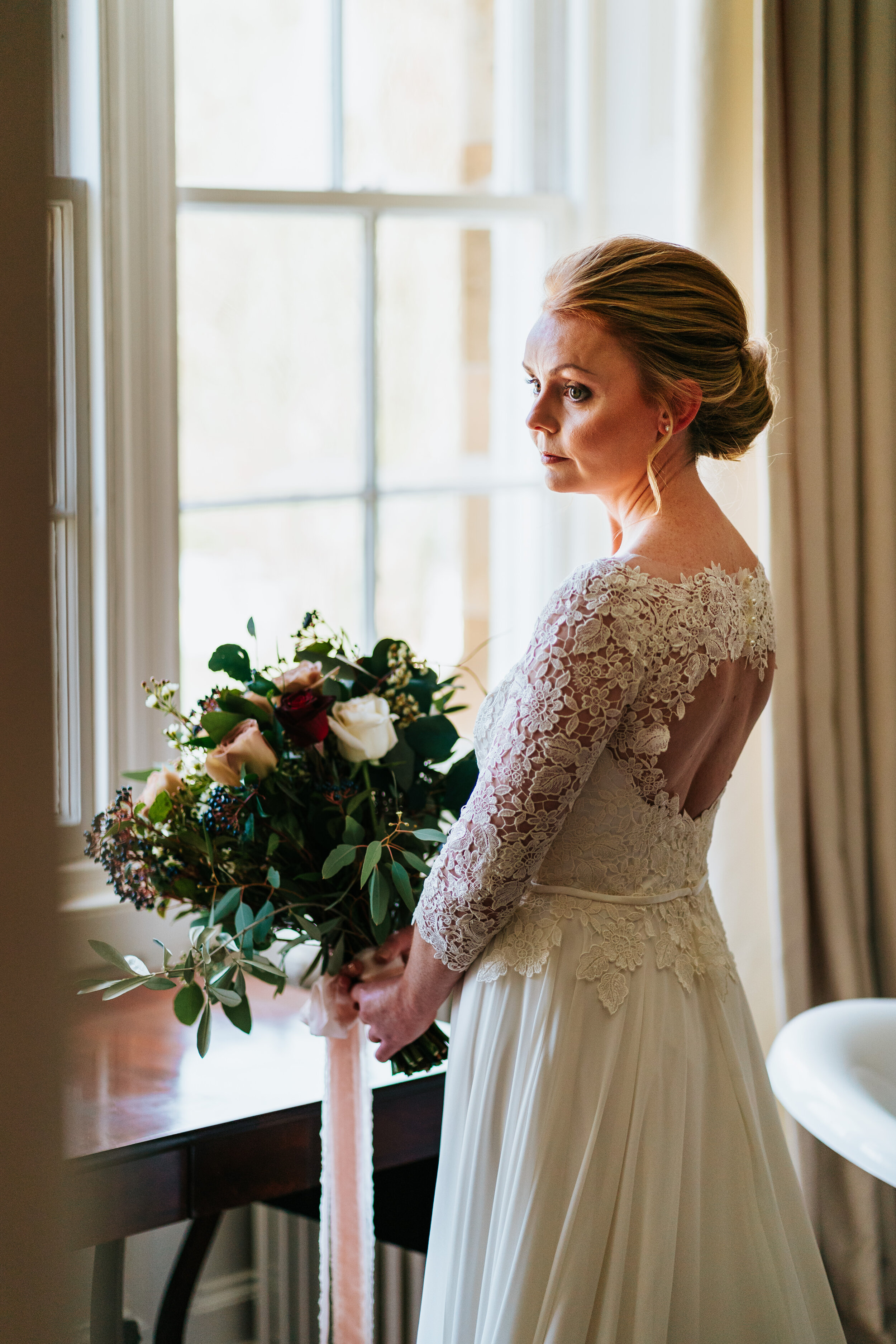 Bride in lace-topped wedding gown standing at window holding bridal bouquet