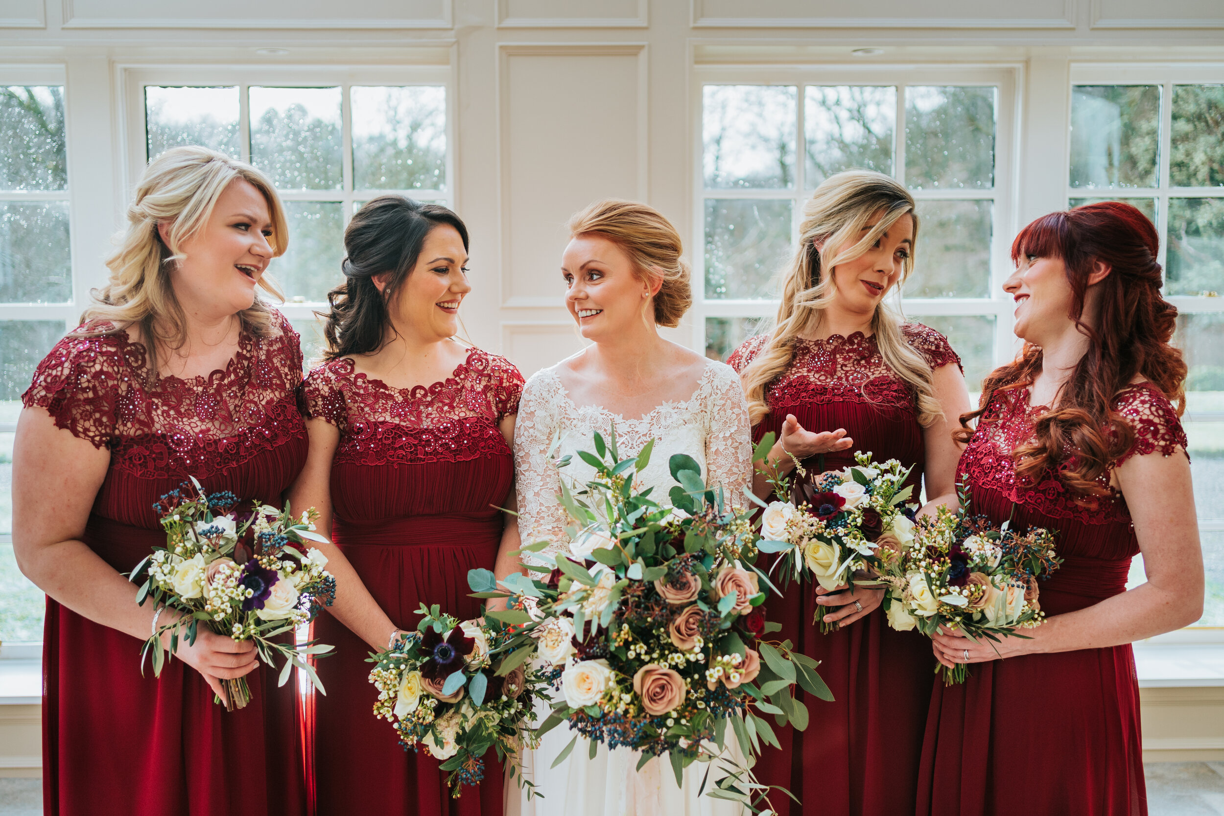 Brides standing with bridesmaids in dark red gowns laughing and holding wedding flowers 