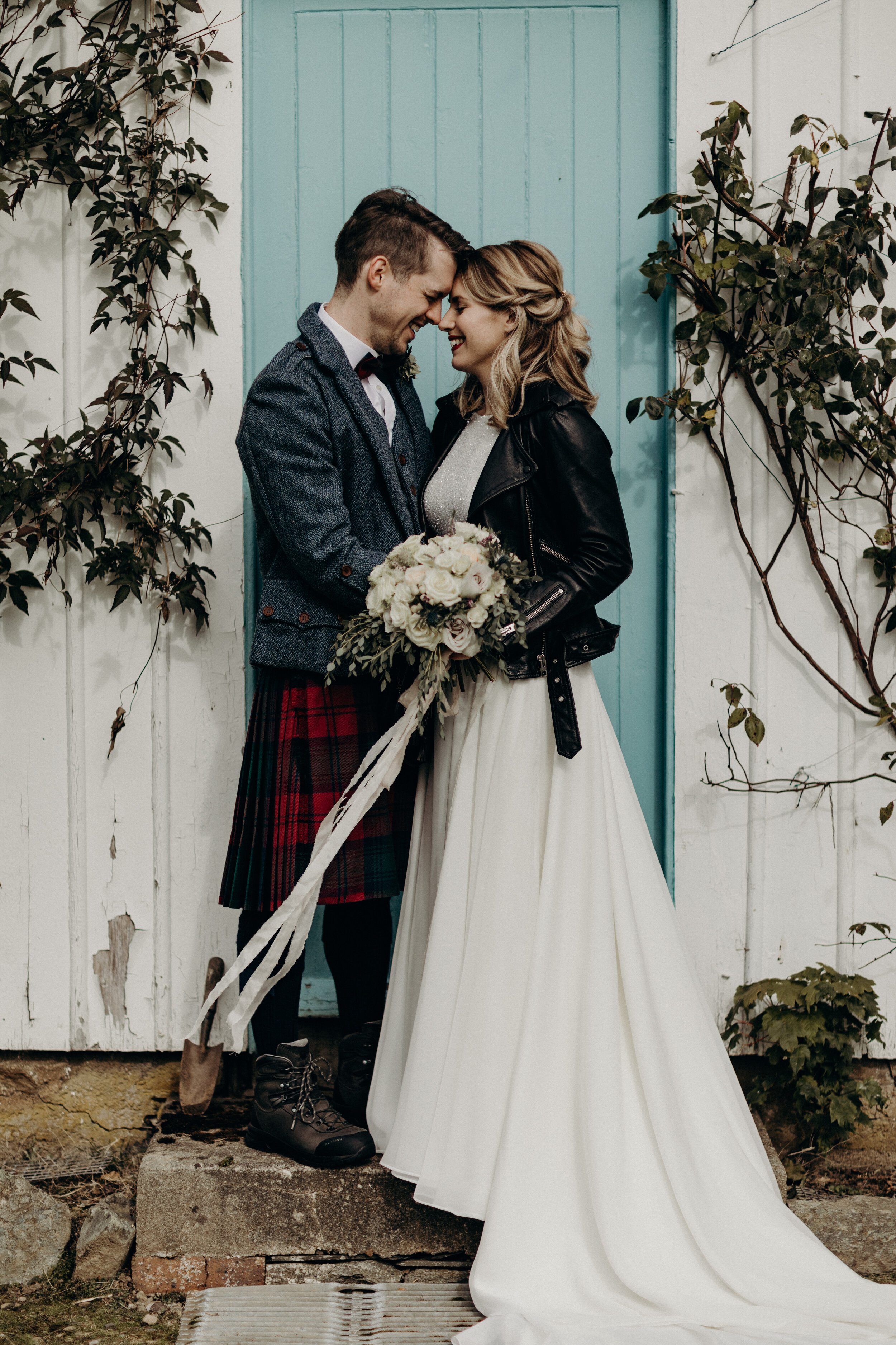 Bride in leather jacket and wedding gown holding bouquet of white roses in Dunkeld