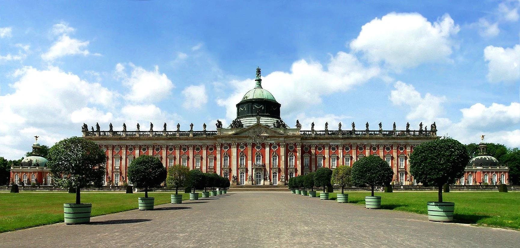 Front view of a grand historic palace with a large central dome, pink walls, white accents, surrounded by a well-maintained lawn with rows of trimmed trees in planters, and a blue sky with some clouds.
