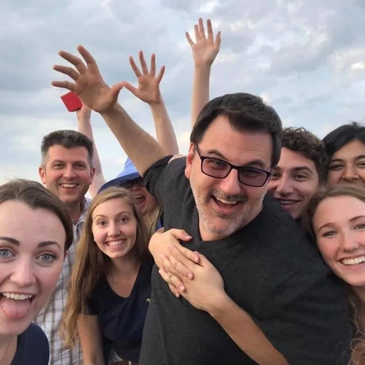 A group of smiling people taking a selfie outdoors, with some raising their hands in the air.