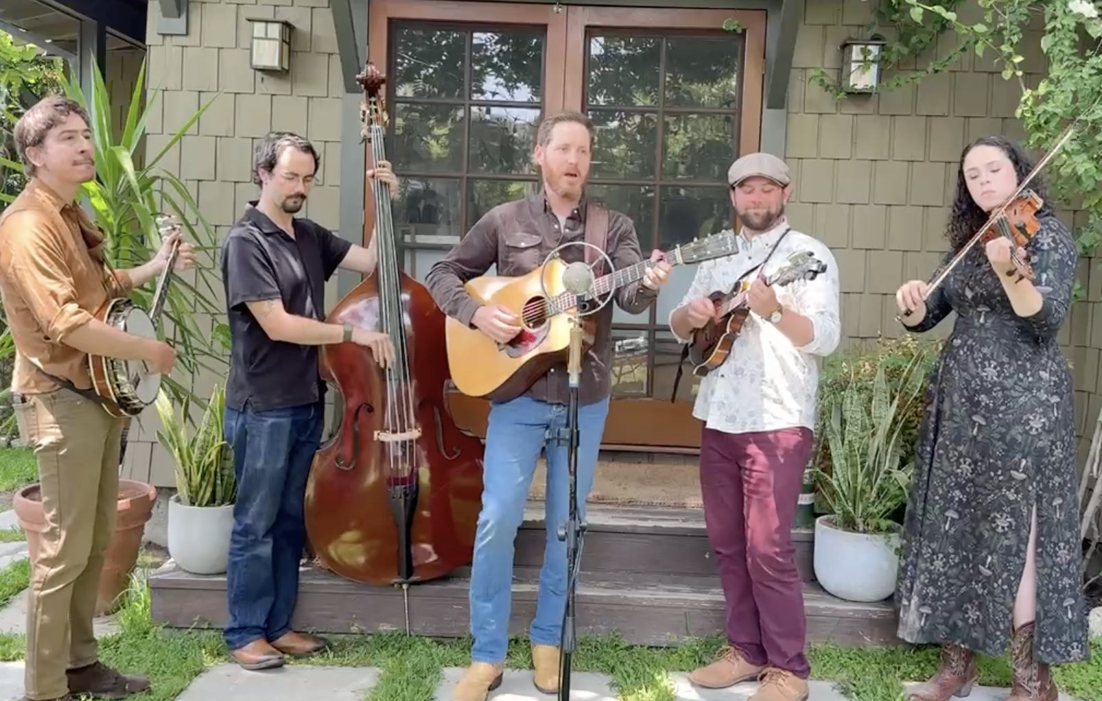 A five-piece band playing string and percussion instruments outdoors in front of a house.