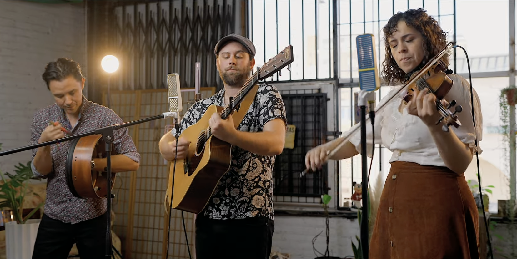 Three musicians performing indoors: a man with a tambourine, a man playing an acoustic guitar, and a woman playing a violin, standing in front of a large window with plants around.