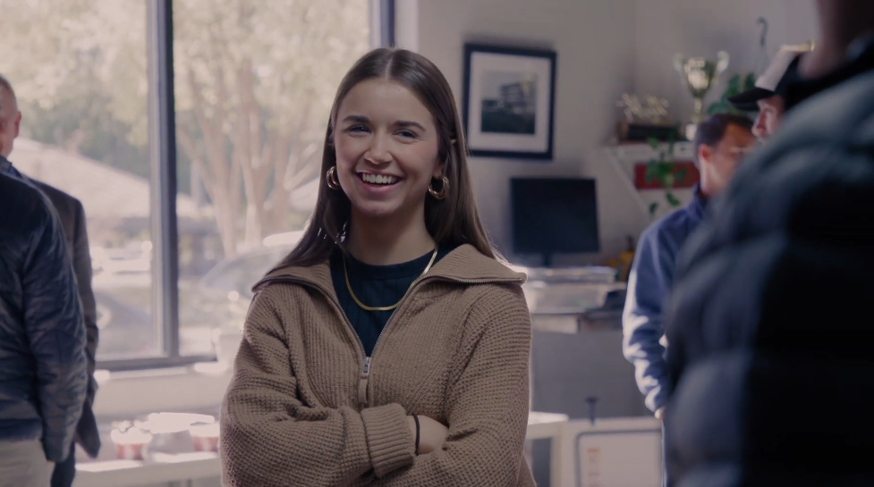 A Chattanooga Fellow with long brown hair, smiling with her arms crossed, wearing gold hoop earrings, a black shirt, and a beige zip-up jacket, standing inside an office environment.