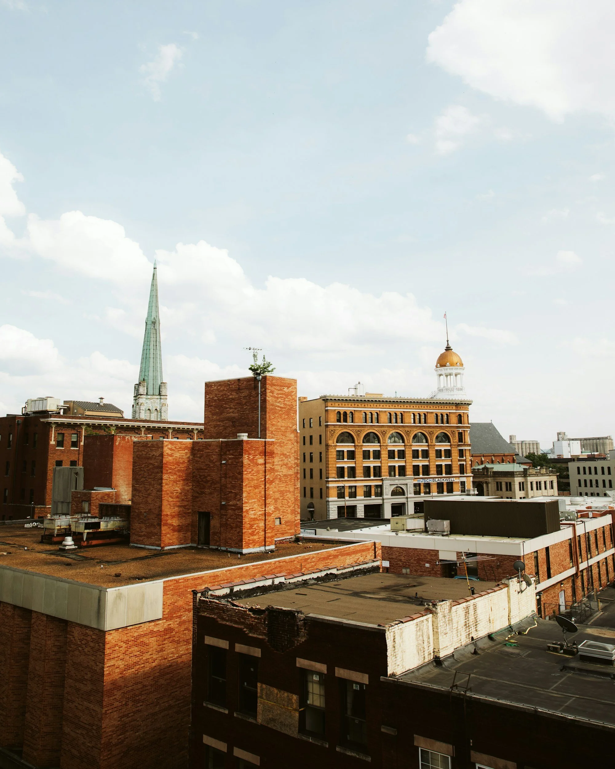 Cityscape with various brick and stone buildings, a church steeple, and a building with a clock tower under a partly cloudy sky.