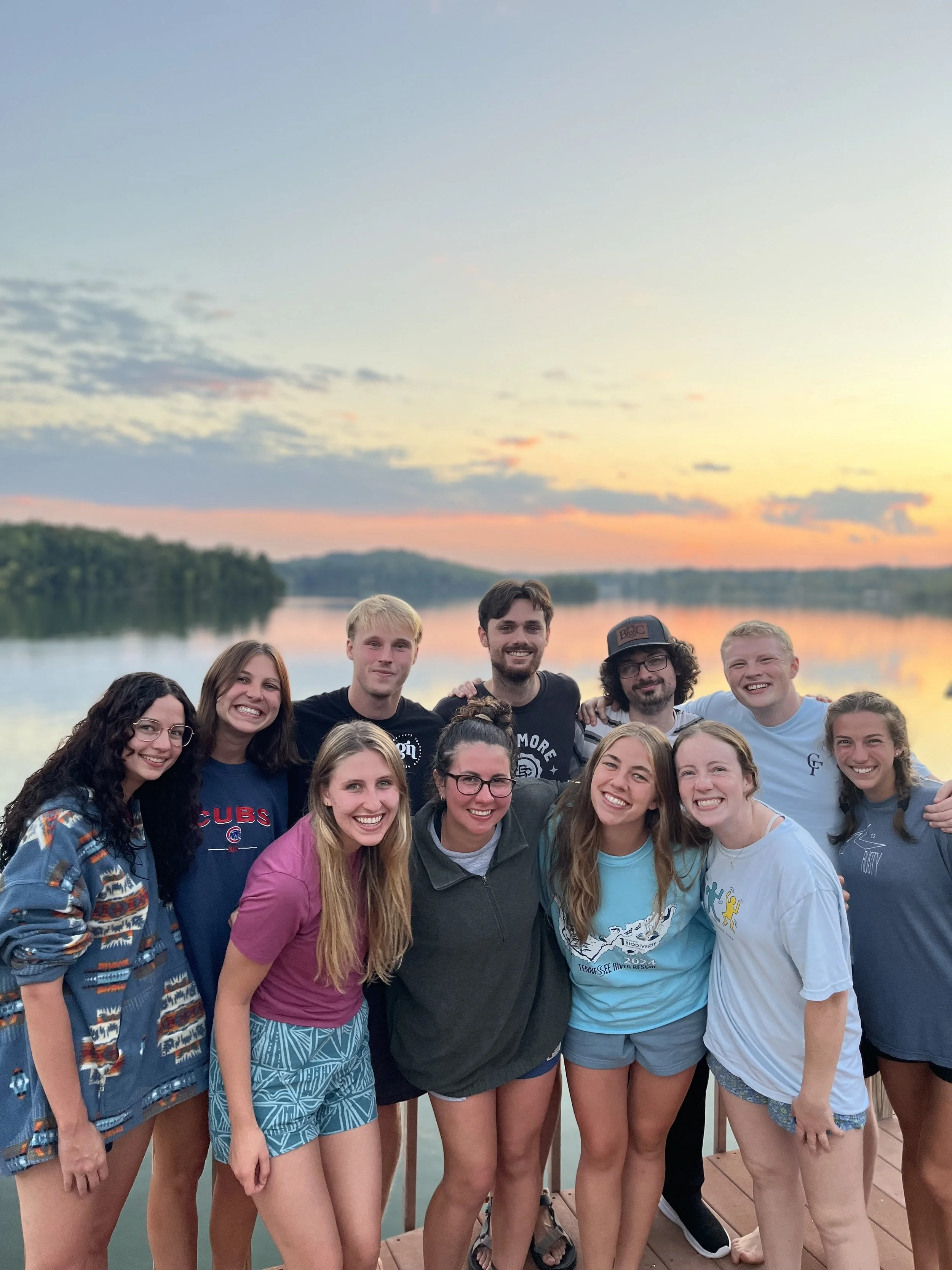 A group of eleven Chattanooga Fellows posing together outdoors during sunset near a body of water, smiling and standing close together.