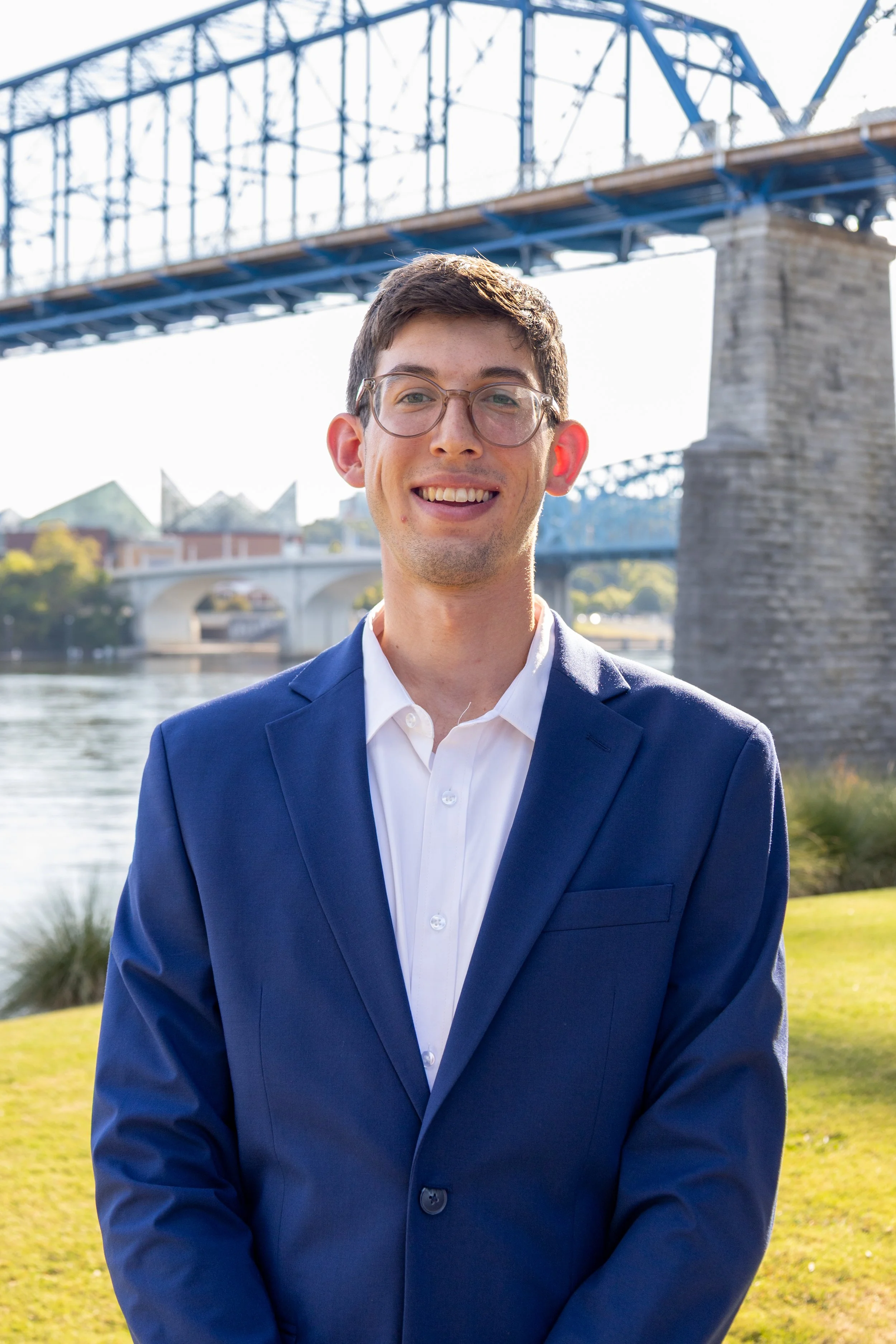 Young man in a suit and glasses, smiling outdoors with blurred trees and a bridge in the background.