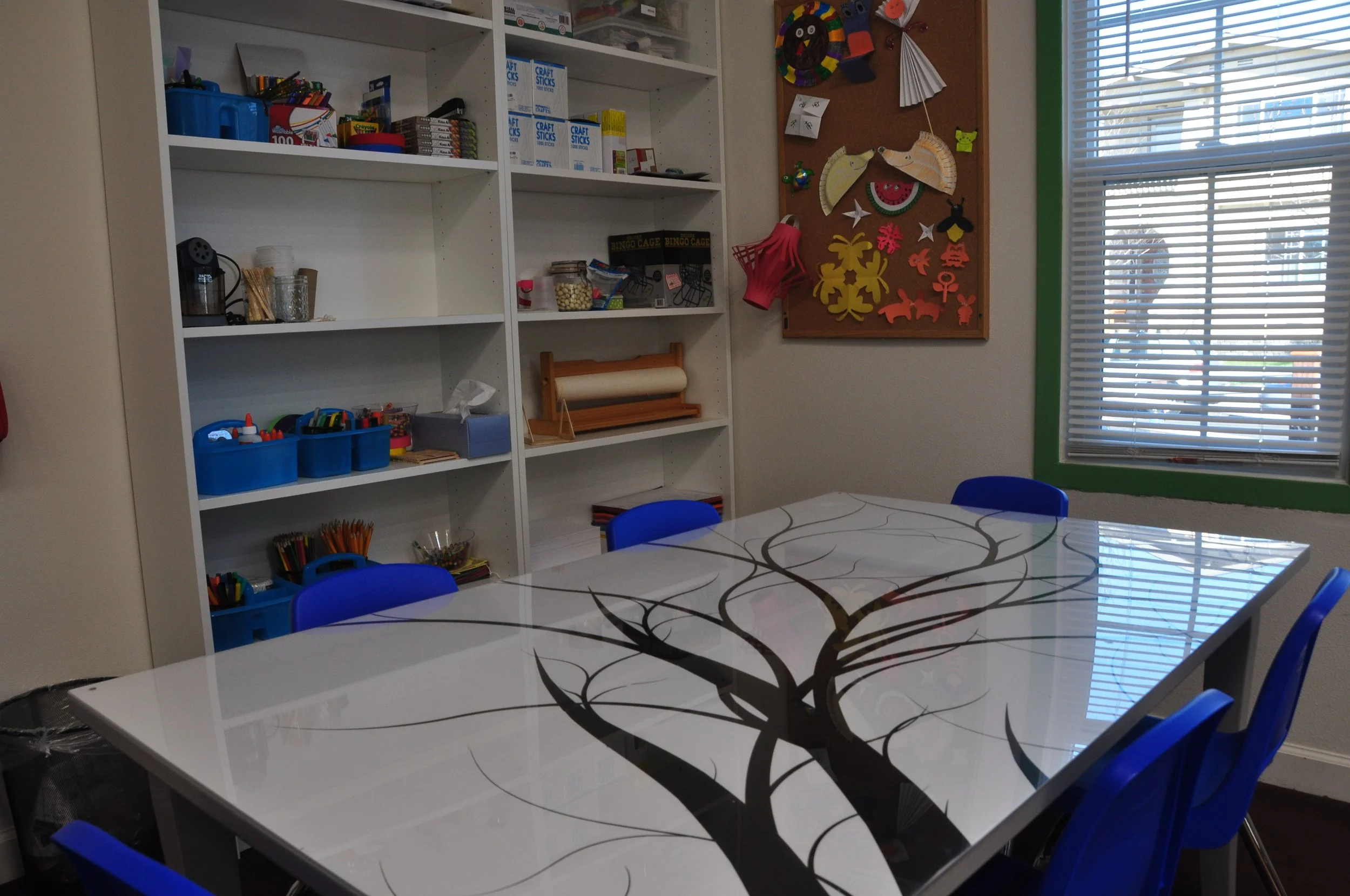 A classroom with a white table featuring a black tree design on the surface, surrounded by blue chairs. In the background, there are white shelves filled with craft supplies and a bulletin board decorated with various paper crafts and decorations. A window with blinds is on the far right, allowing light to enter.