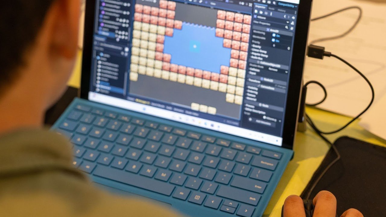 A person working on a laptop with game development software open, showing a pixel art game level with bricks and a sky background.