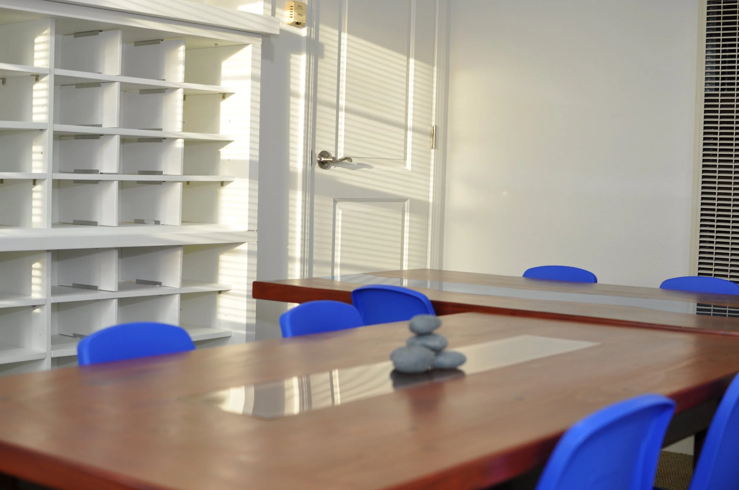 A room with a white door, white shelving unit, blue chairs around a wooden table, and sunlight casting shadows through window blinds.