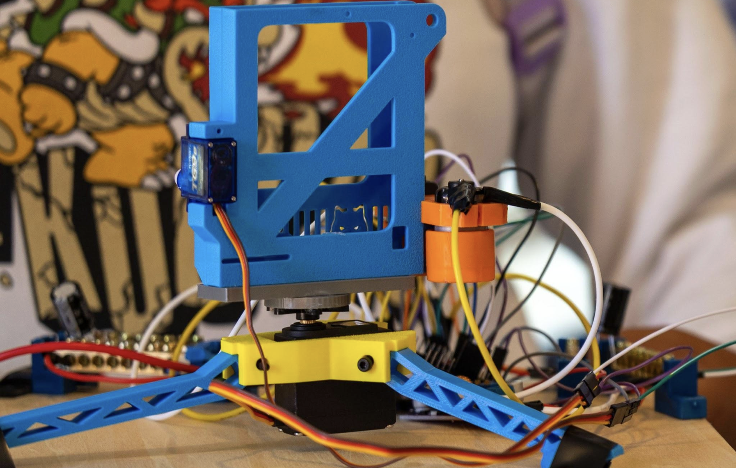 A close-up of a blue 3D-printed robotic structure with wires and electronic components, including a small camera or sensor attached, set on a wooden surface with a colorful cartoon mural in the background.