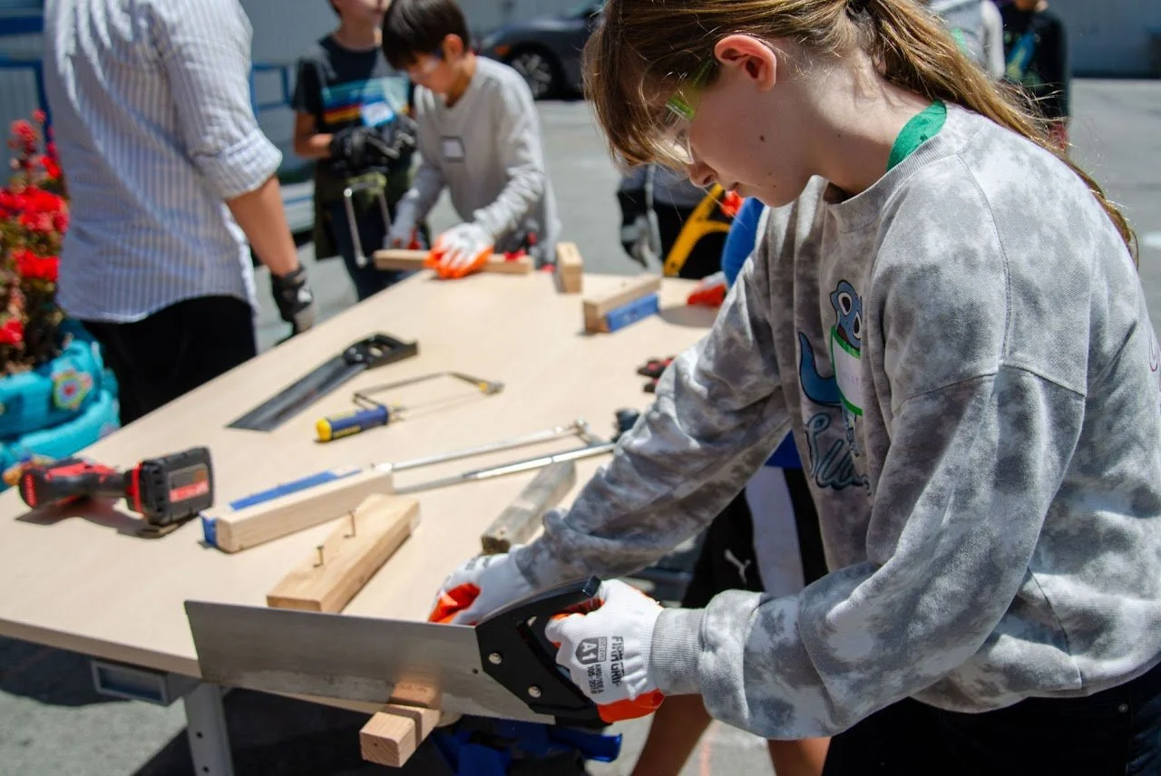A girl wearing safety glasses and gloves using a handsaw to cut a piece of wood on a workbench. Other children and an adult are working on a woodworking project outdoors.