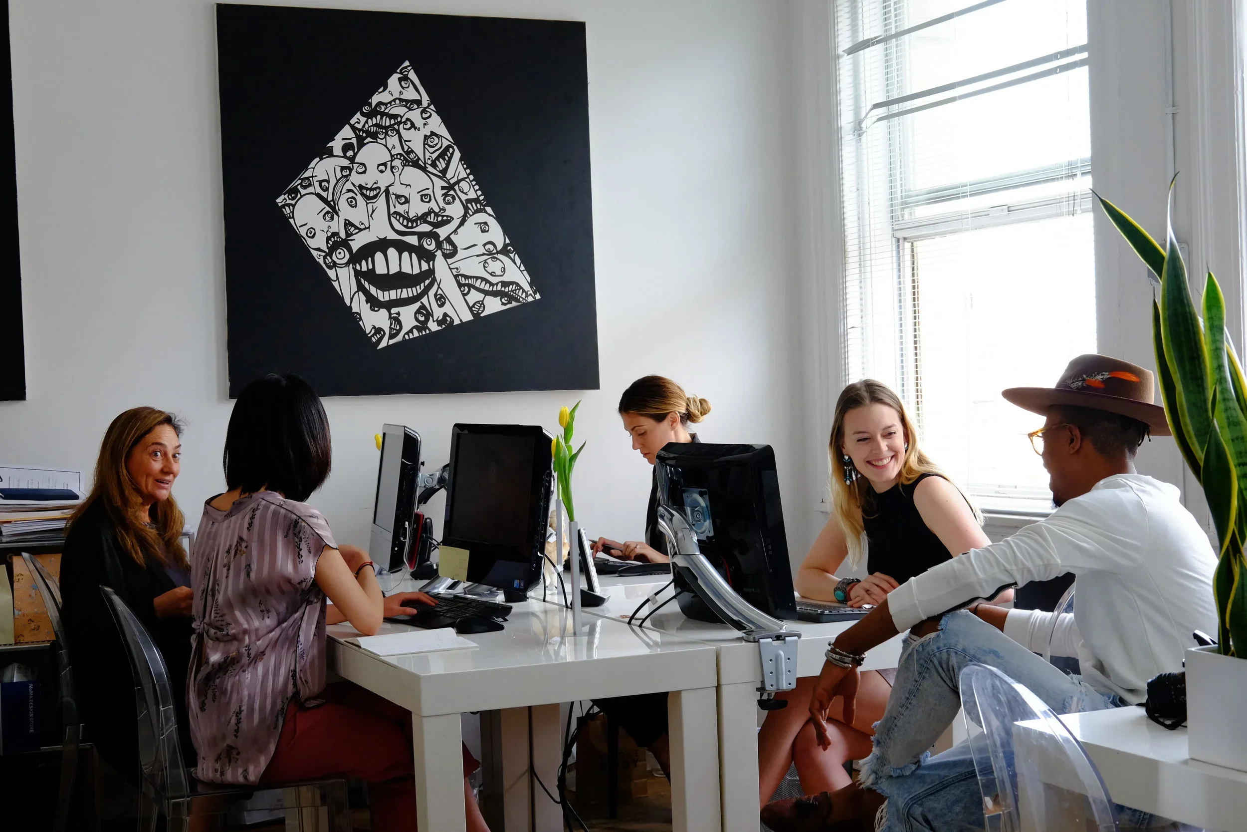 Groupe de cinq personnes assises autour d'une table de bureau avec des ordinateurs, dans une salle lumineuse avec des fenêtres, discutant et souriant.