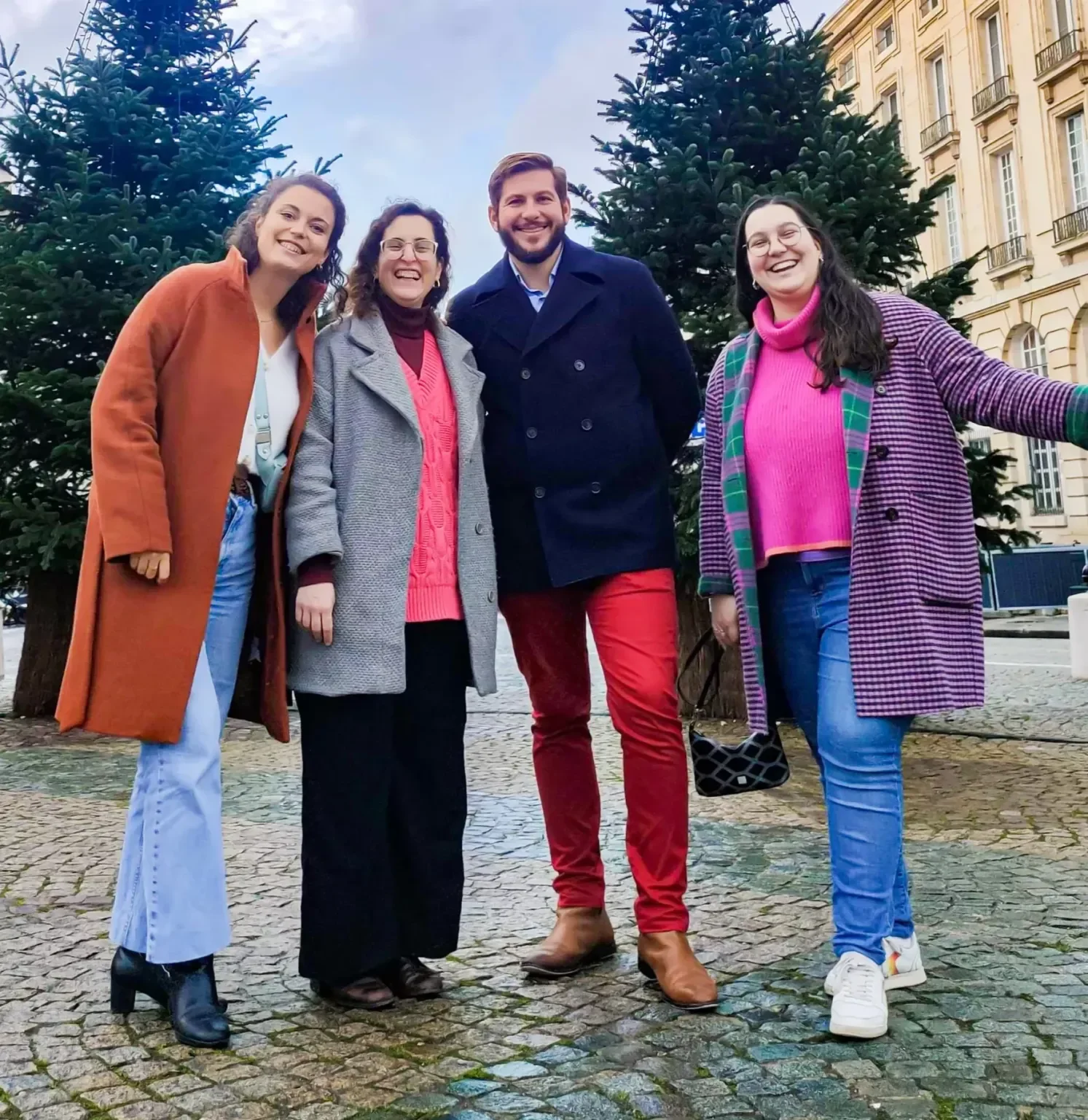 Groupe de quatre personnes souriantes posant devant deux sapins de Noël, en ville, pendant la journée.