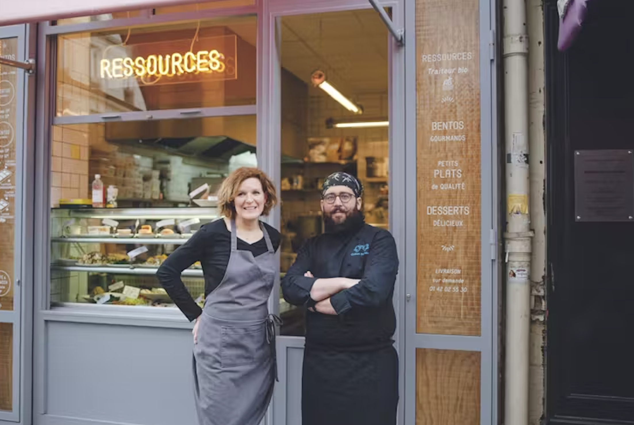 Deux chefs posent devant la vitrine d'une boutique de restauration à Paris, avec un néon 'Ressources' au-dessus d'eux, et un menu affiché à droite.