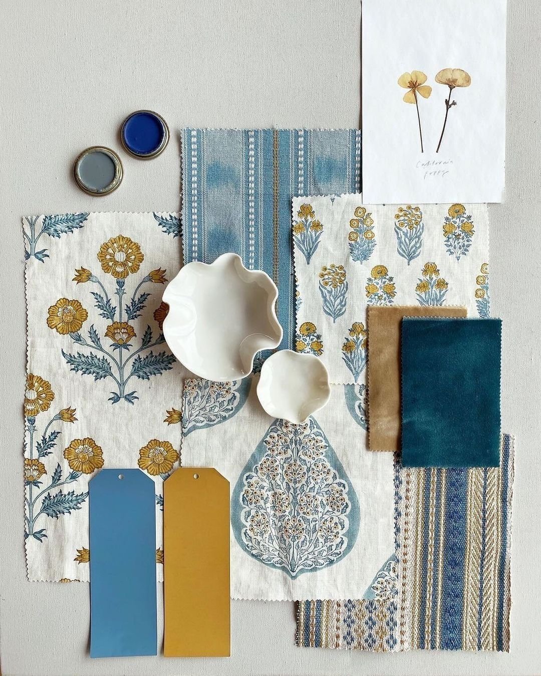 Échantillons de tissus bleus et beiges avec motifs floraux, deux petits bols blancs, deux pots de peinture bleue et grise, une photo de fleurs séchées, et deux étiquettes colorées jaunes et bleues.
