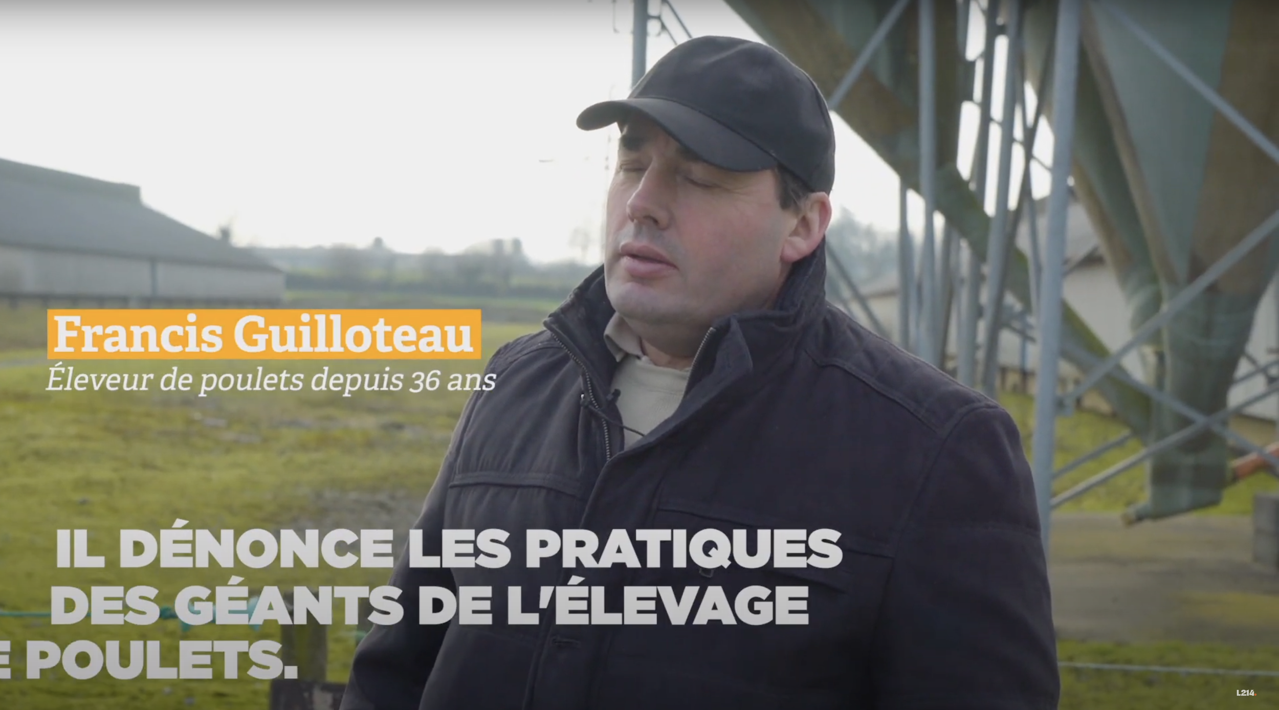 Un homme portant une casquette noire et une veste noire, avec des yeux fermés, dans un champ avec une structure métallique en arrière-plan.