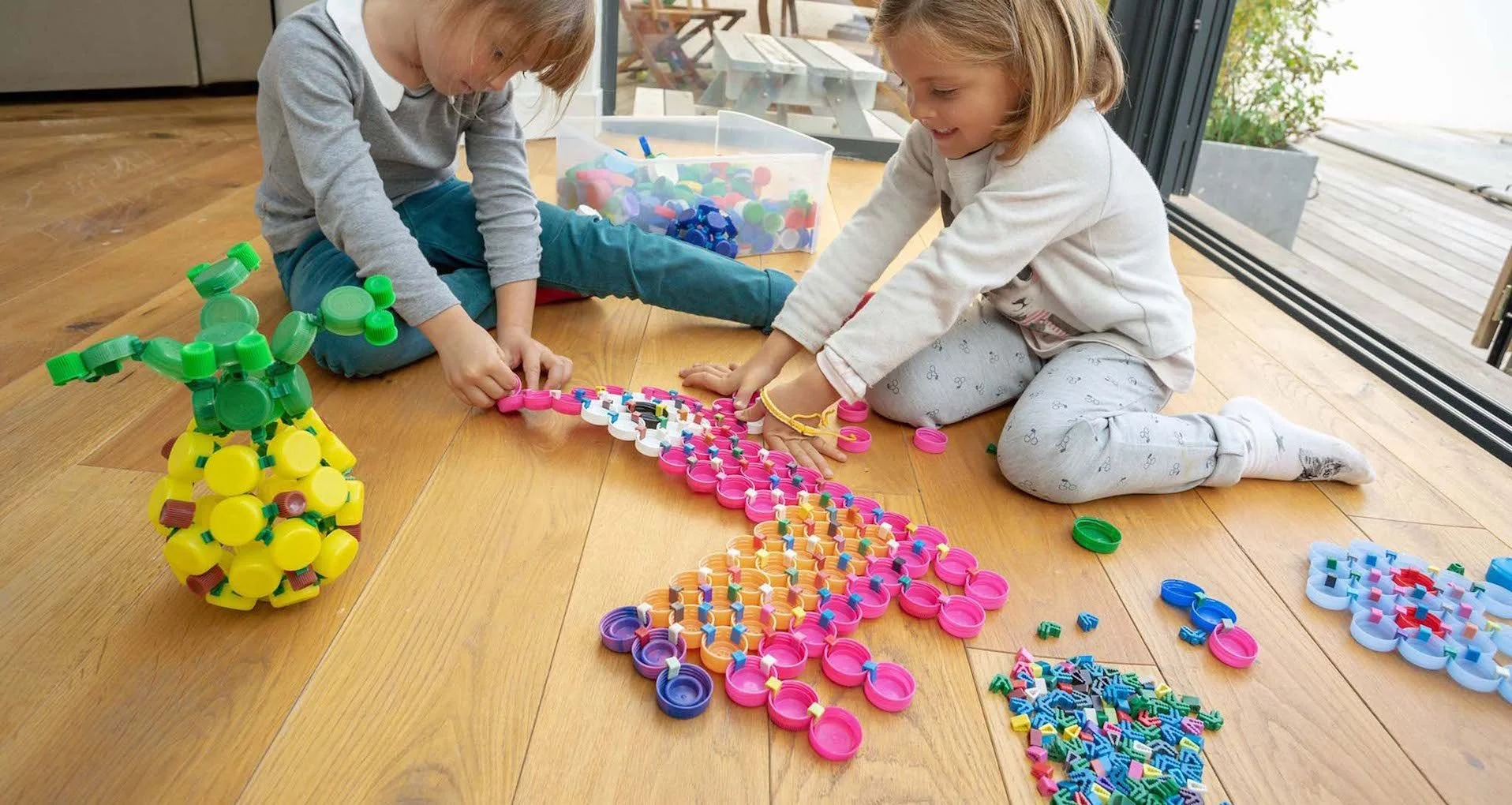 Deux enfants jouant avec des jouets de construction en plastique sur un sol en bois, entourés de différentes pièces colorées et d'une grande figure en forme de bouquet de fleurs.