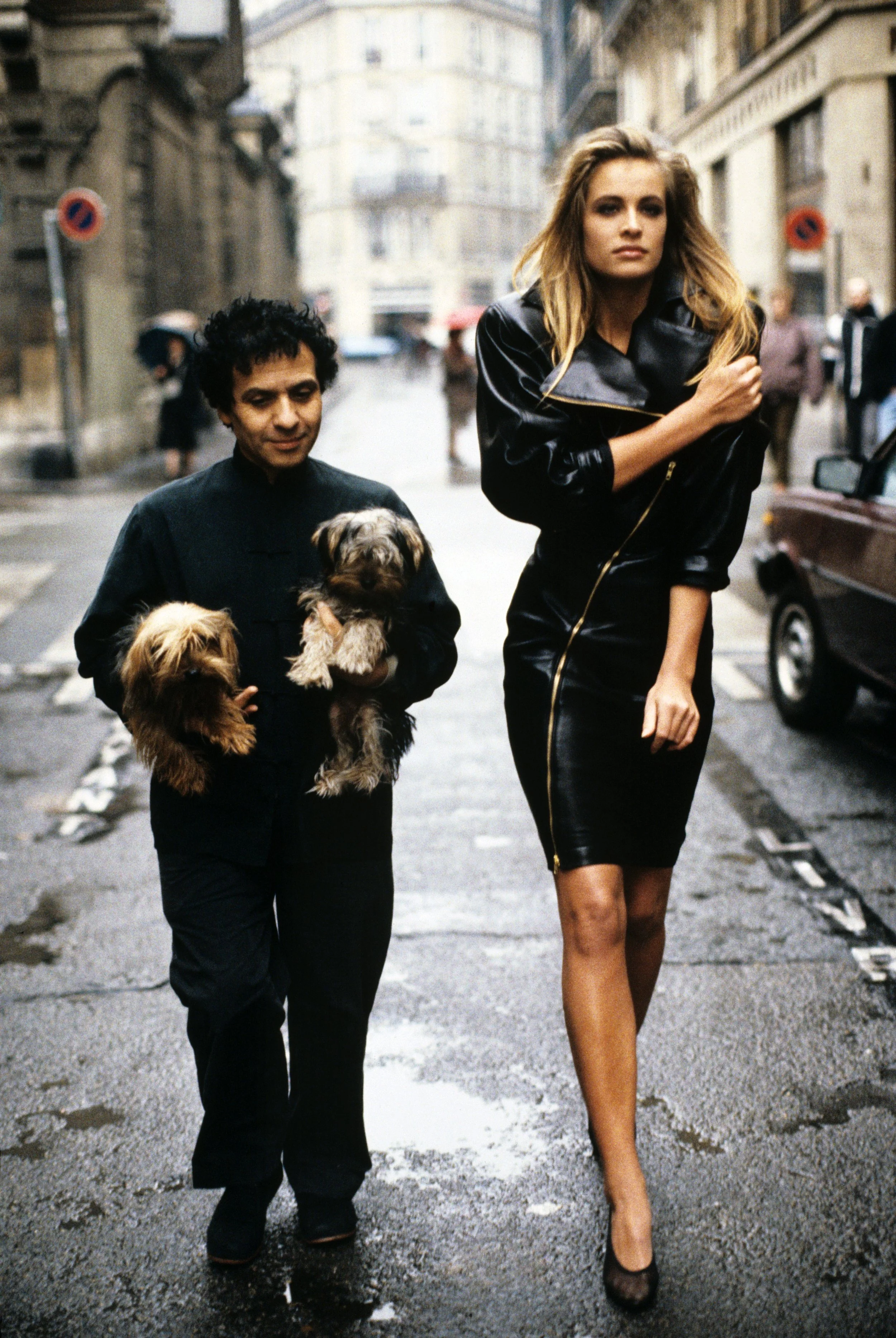Fashion designer Azzedine Alaïa holding his two Yorkshire terriers, Patapouf and Wabo, walking in Paris street with model Frederique who wears one of his creations, a black leather zippered dress, 1986. Photograph Courtesy of Arthur Elgort.