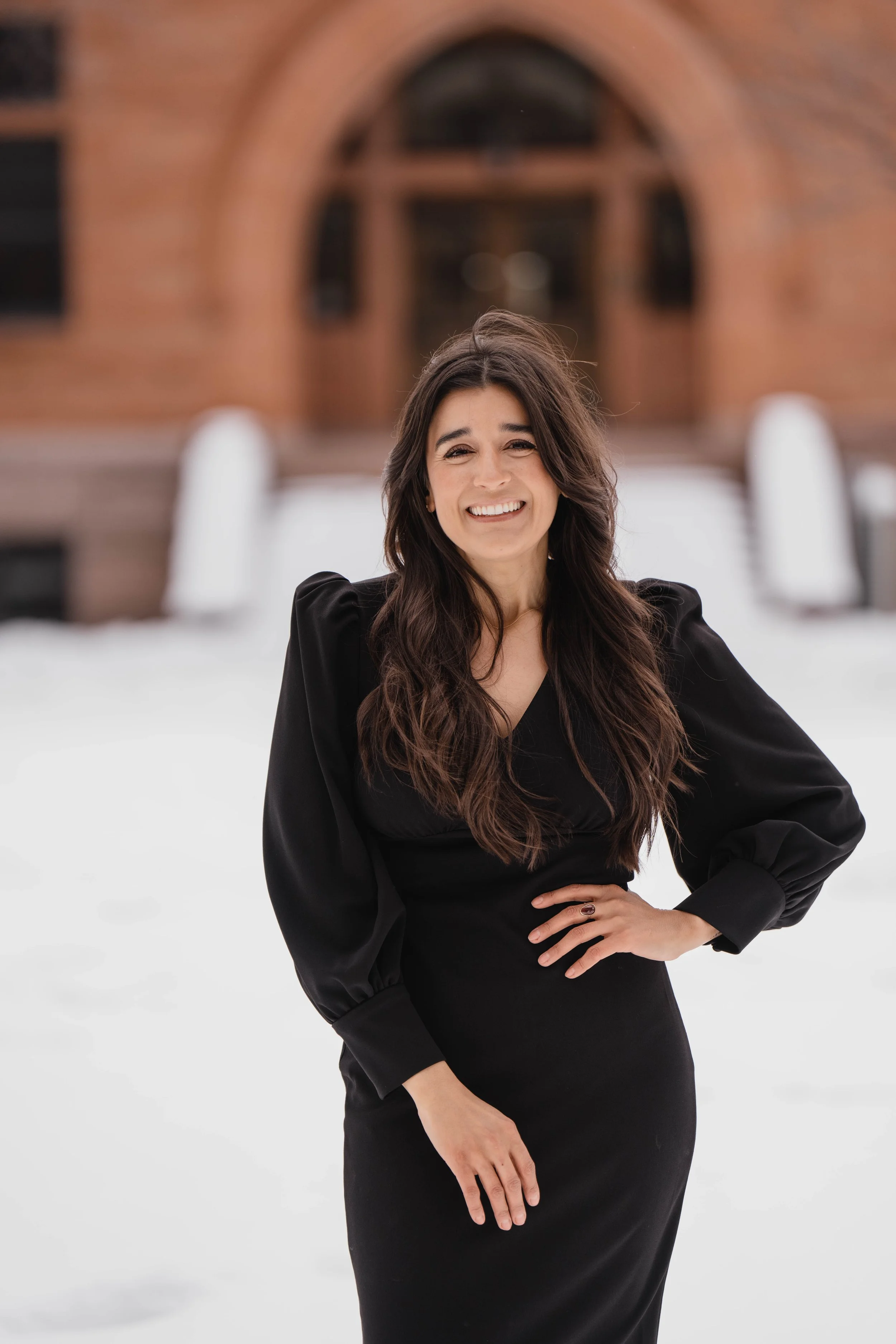 A woman in a black dress standing outdoors in front of a brick building, smiling with one hand on her hip.