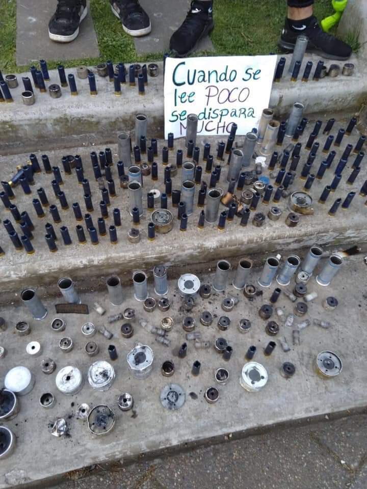  A selection of used shotgun hulls and remnants of tear gas canisters.  The sign reads (roughly) “Those who read little, shoot much.” 