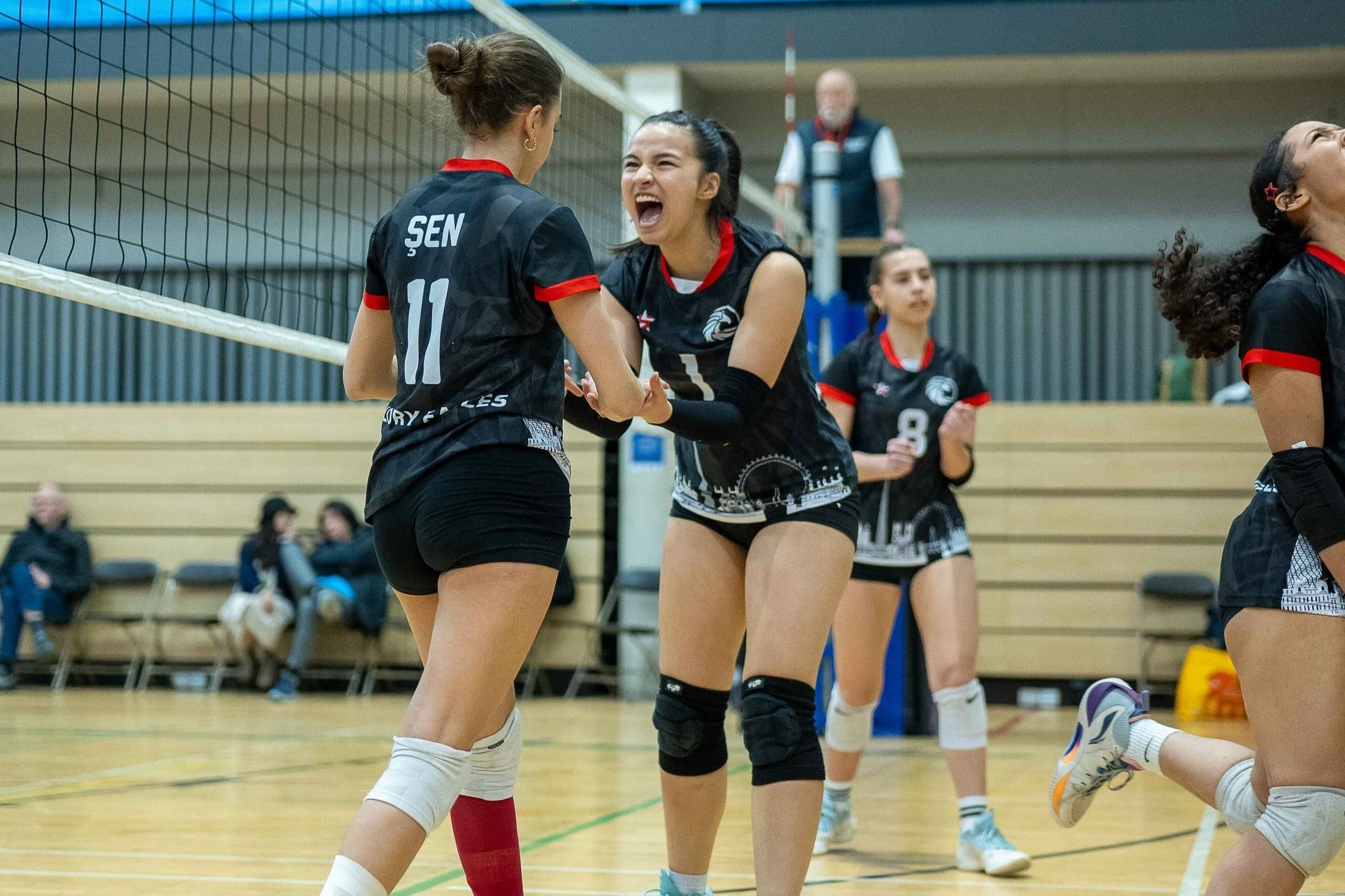 Four female volleyball players in black uniforms celebrating on an indoor court, with spectators seated in the background.