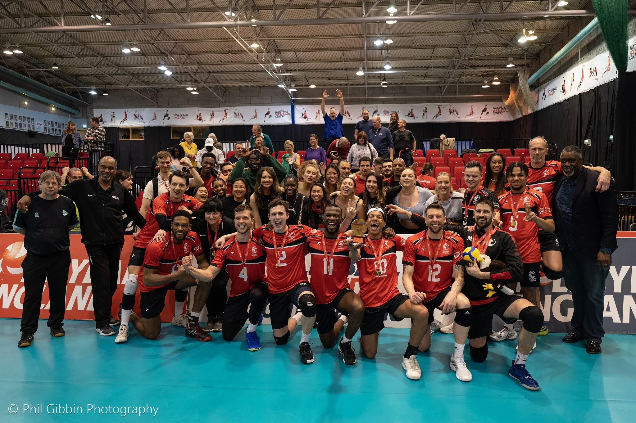 A group of volleyball players and supporters celebrating their victory on an indoor court, with medals around their necks and a trophy. They are smiling, kneeling and standing with arms around each other, some holding plush toys and volleyballs. Behind them are empty red seats and a background banner.