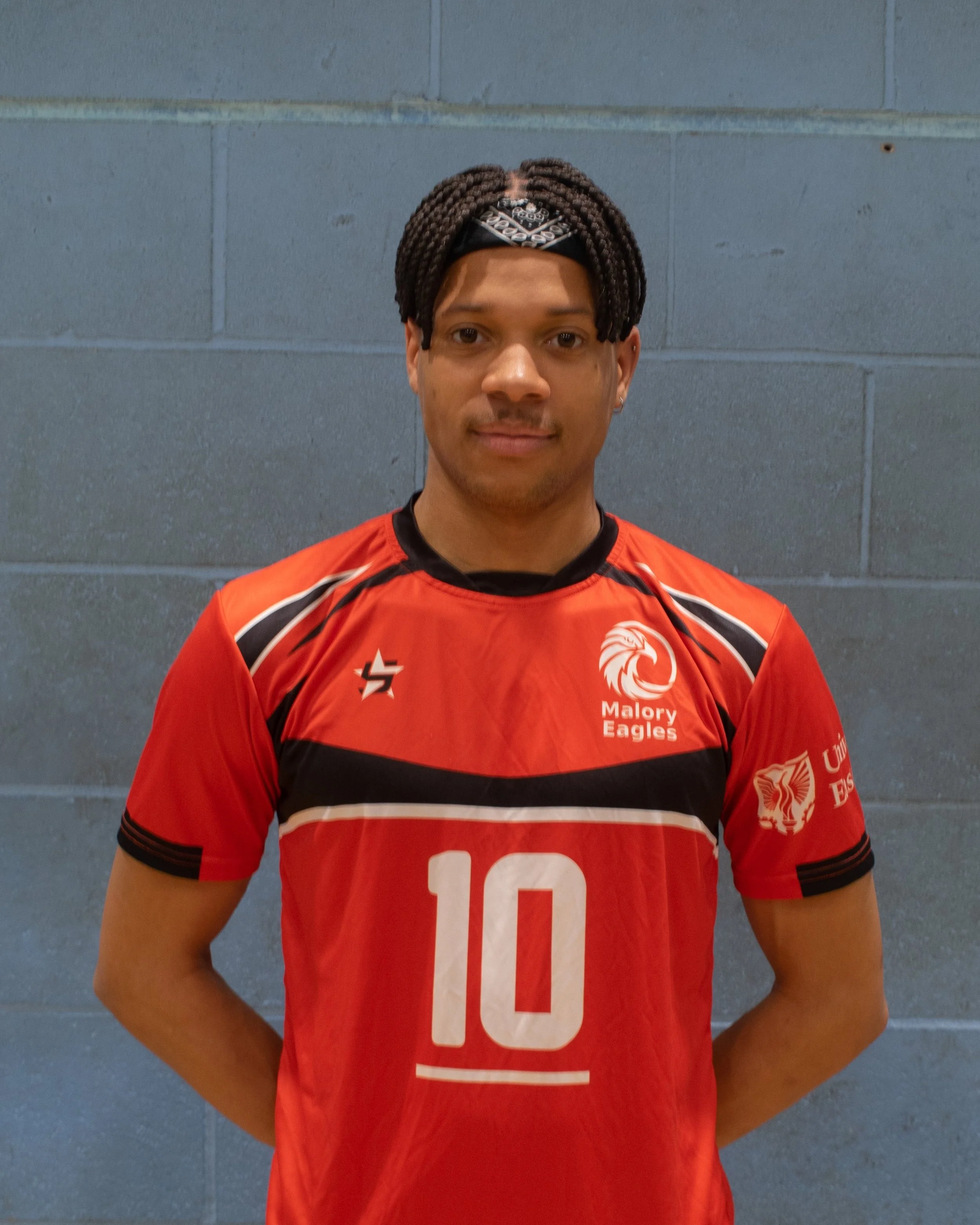 A young man wearing a red volleyball jersey with the number 10 and team logos, standing against a blue brick wall.
