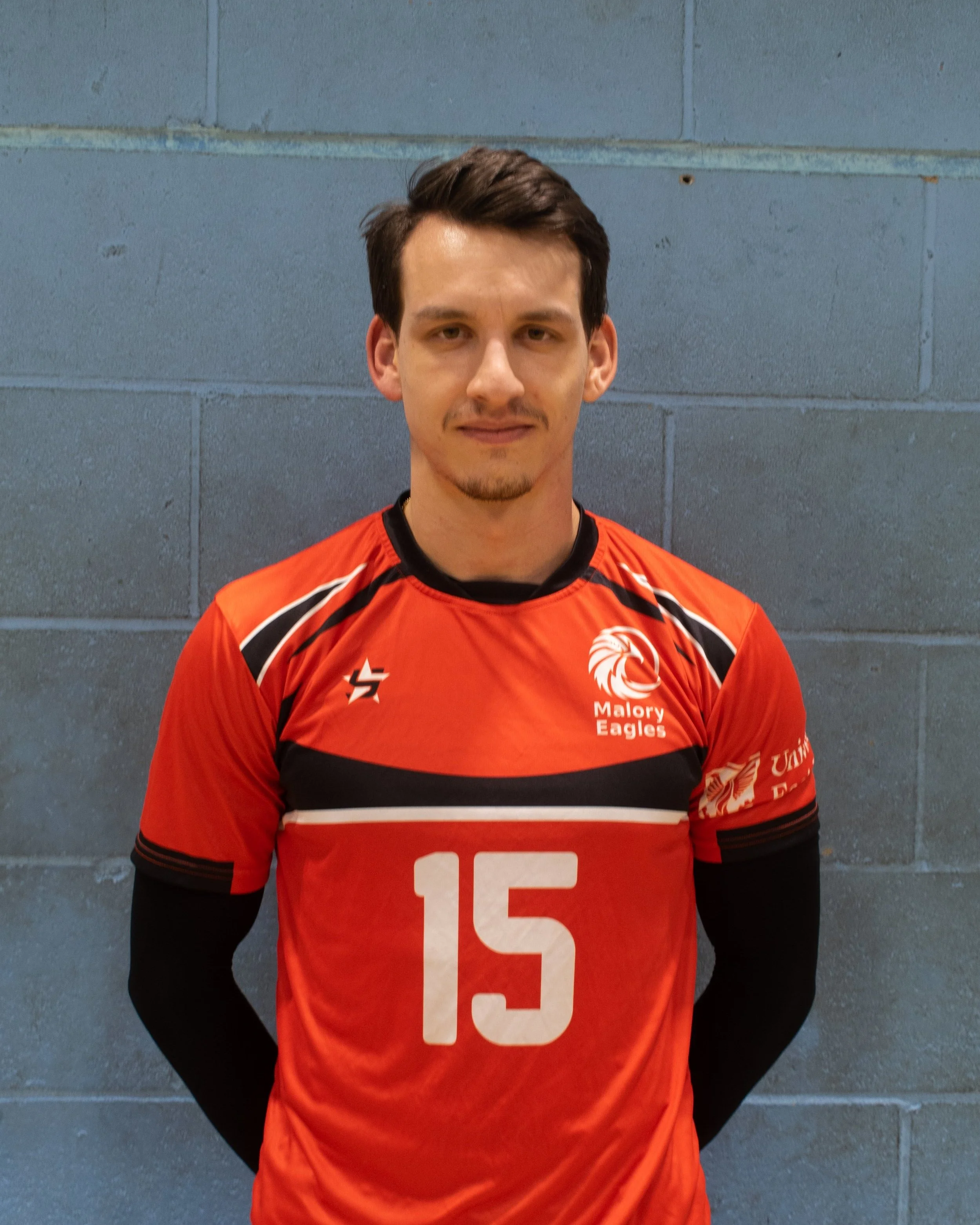 A young man in a red sports jersey with the number 15, standing against a gray brick wall.