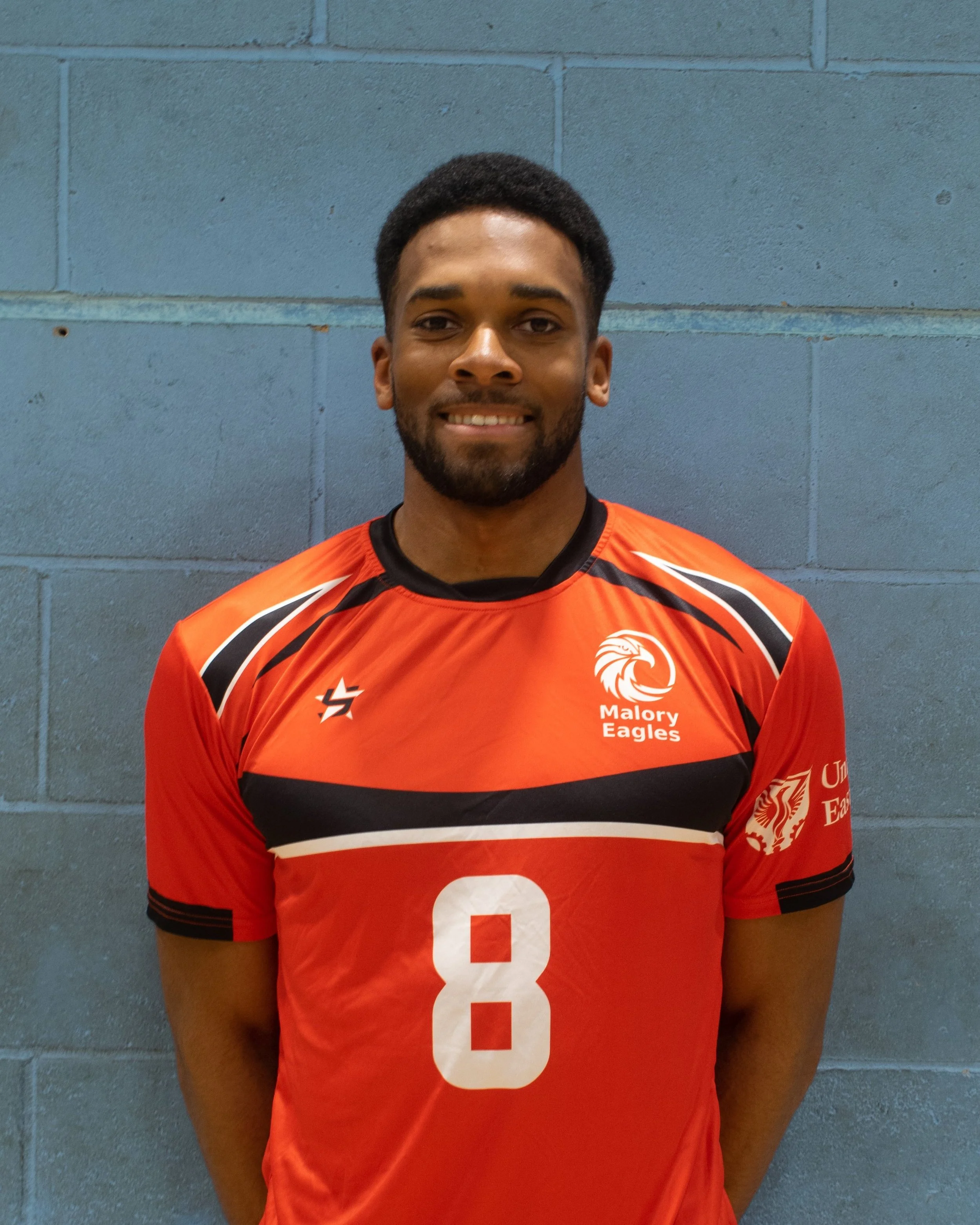 A young man smiling, wearing a red sports jersey with the number 8, standing against a blue brick wall.