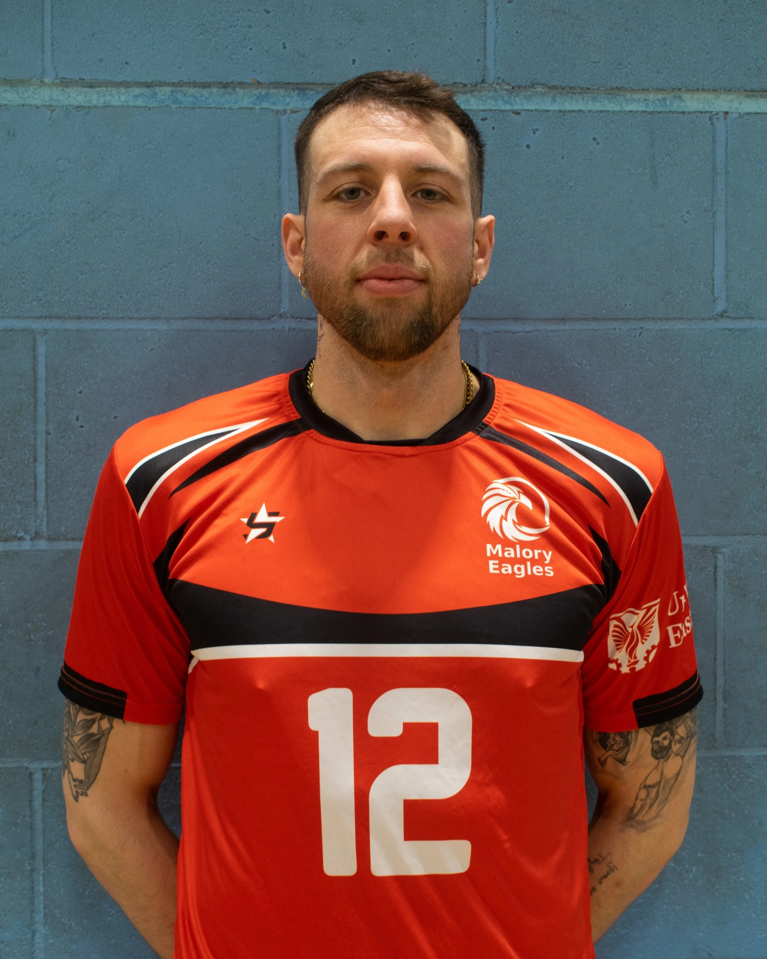 A young man with short brown hair, a beard, and tattoos on his arms, wearing an orange sports jersey with the number 12, the Malory Eagles logo, and various sponsor logos, standing in front of a blue brick wall.